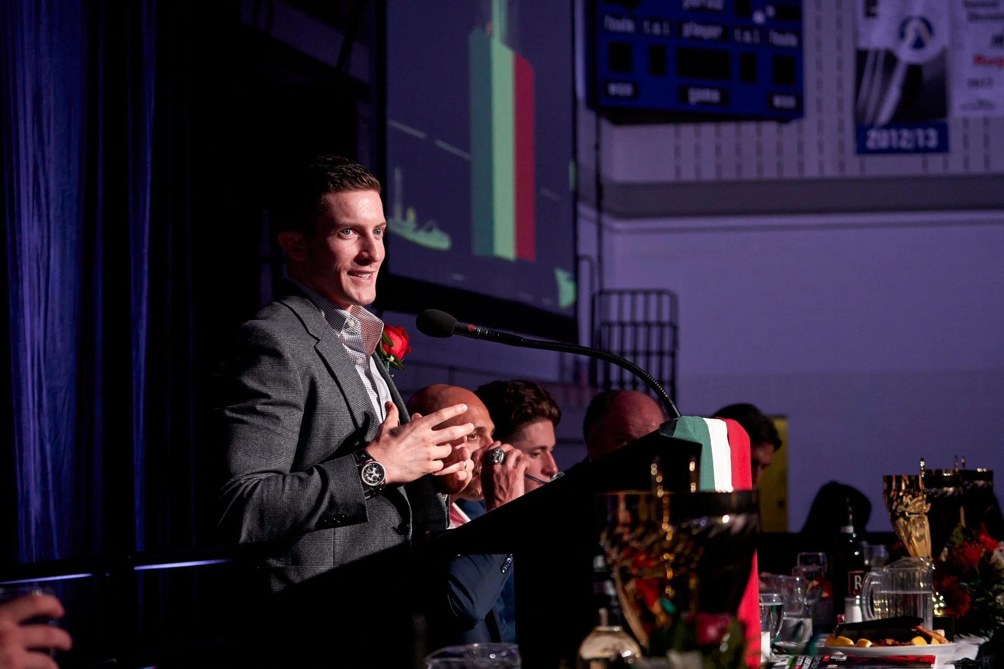 Olympic gymnast Jackson Payne in gray suit with red rose boutonniere addressing the crowd at Calgary Italian Sportsman event – passionate delivery under dramatic lighting. Keynote Speaker captured by Stance Creative.