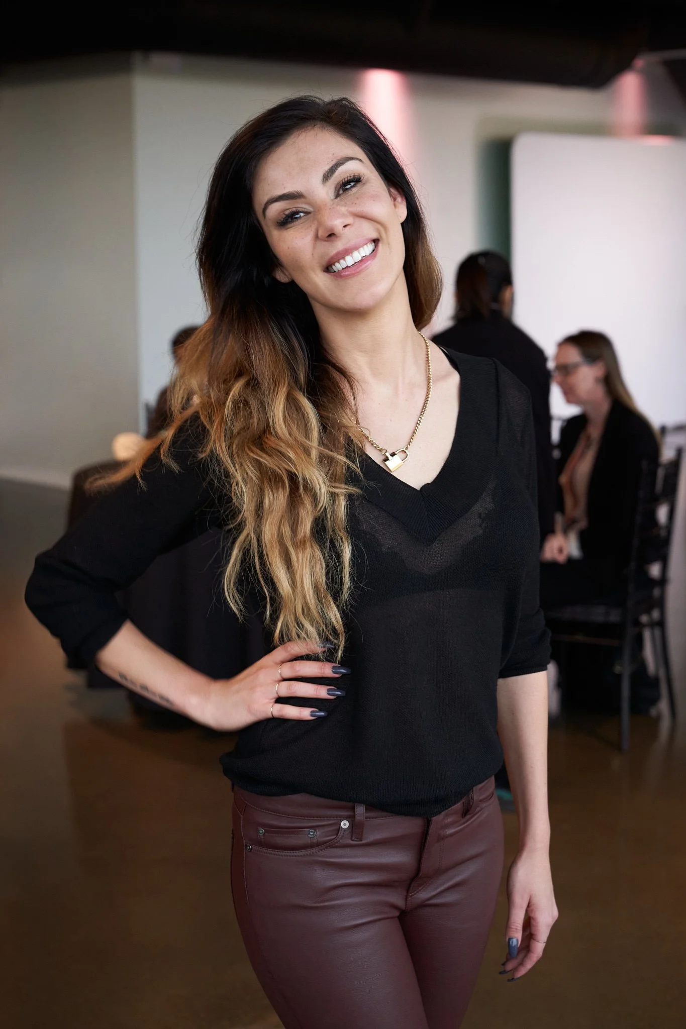 Confident woman in black sheer top and maroon leather pants posing with hand on hip at Calgary networking reception – bold style against an industrial backdrop. Striking portrait captured by Stance Creative event photography. 