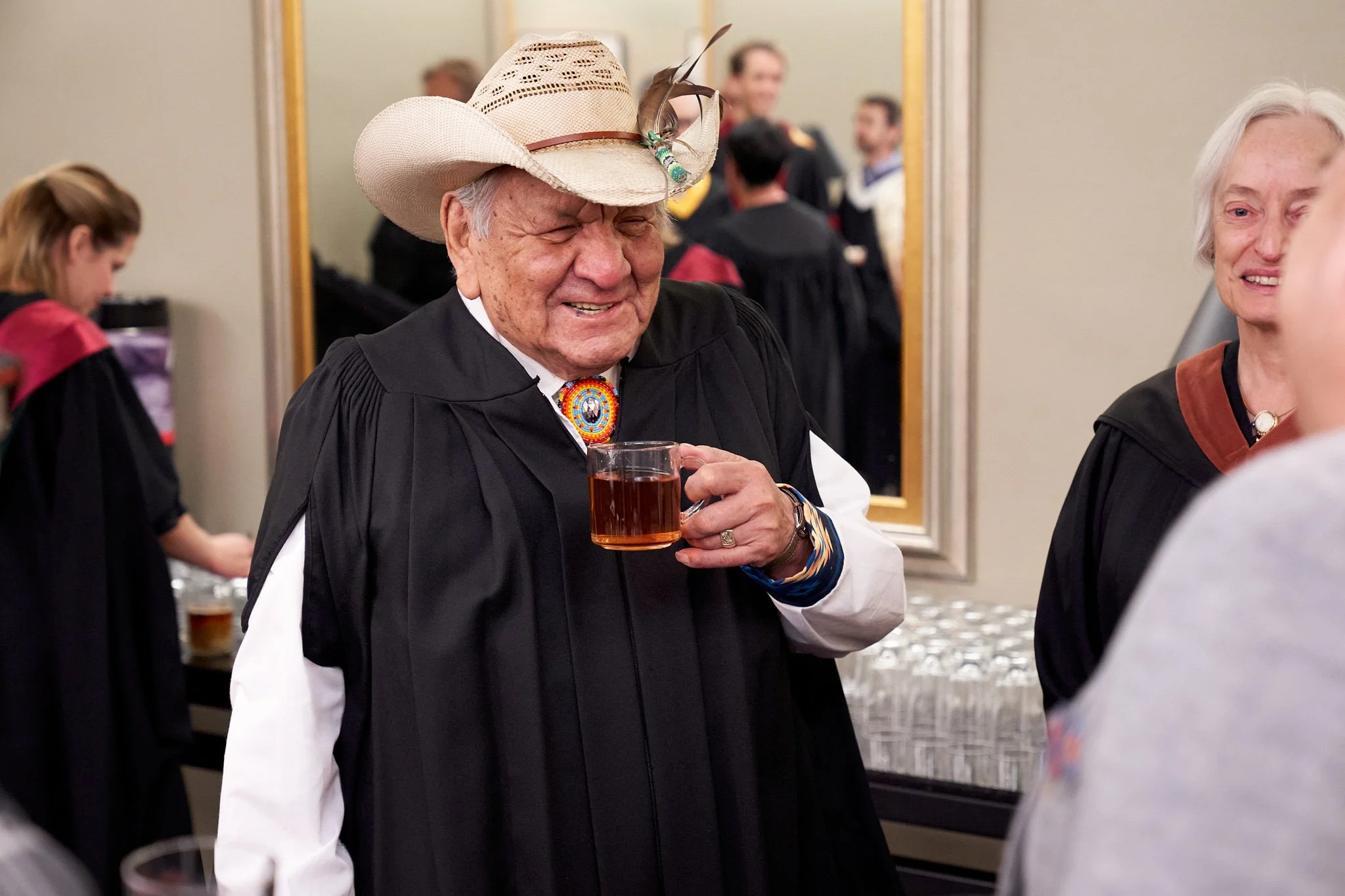 Artist  Alex Janvier in a cowboy hat adorned with feathers and beaded medallion sharing a warm smile moments before receiving his honorary MFA. Authentic portrait by Stance Creative.