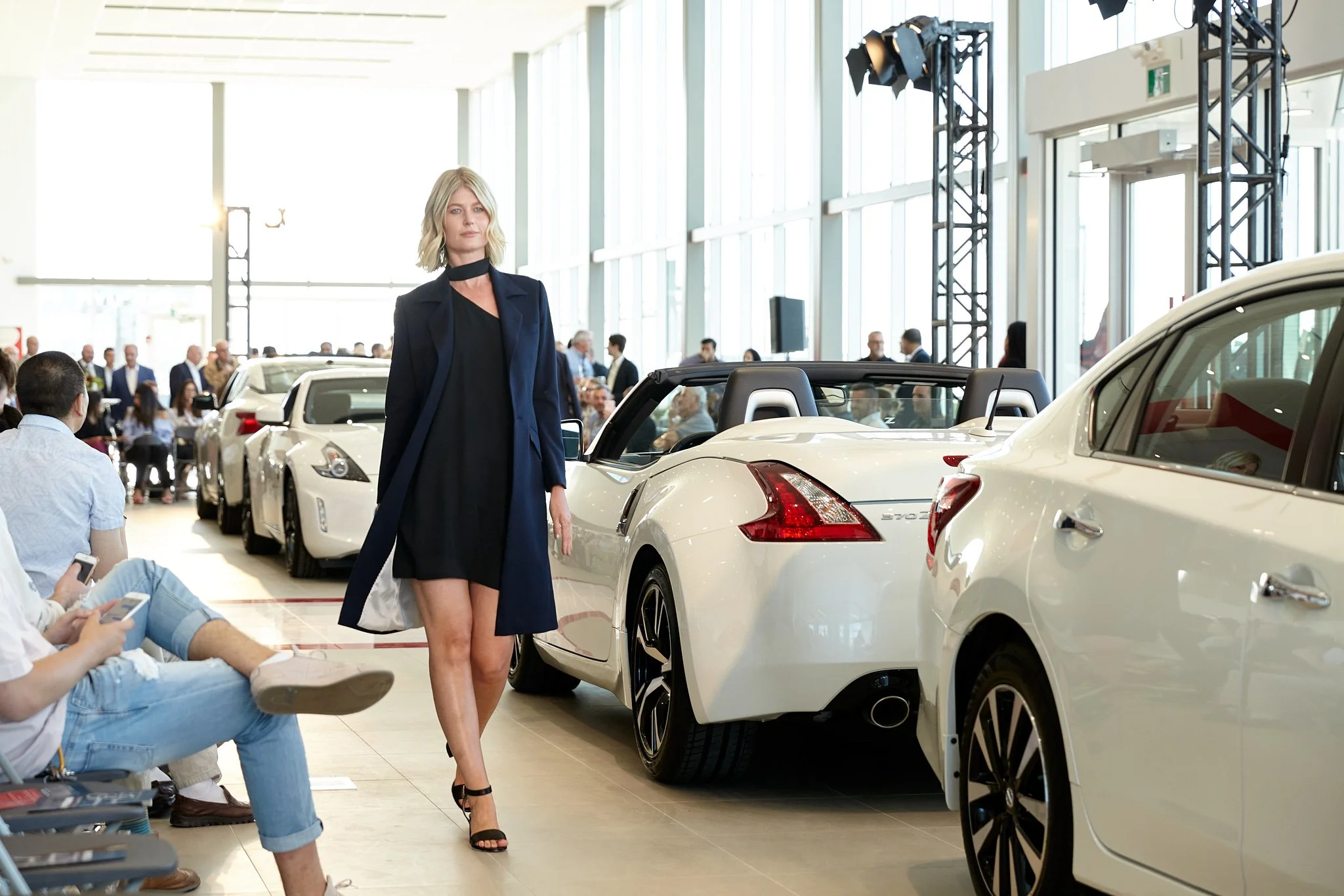 Fashion runway model in navy coat and black dress striding past luxury Nissan cars at the opening of the Calgary Country Hills Nissan  auto showroom event. Captured by Stance Creative, professional Calgary event photographer.