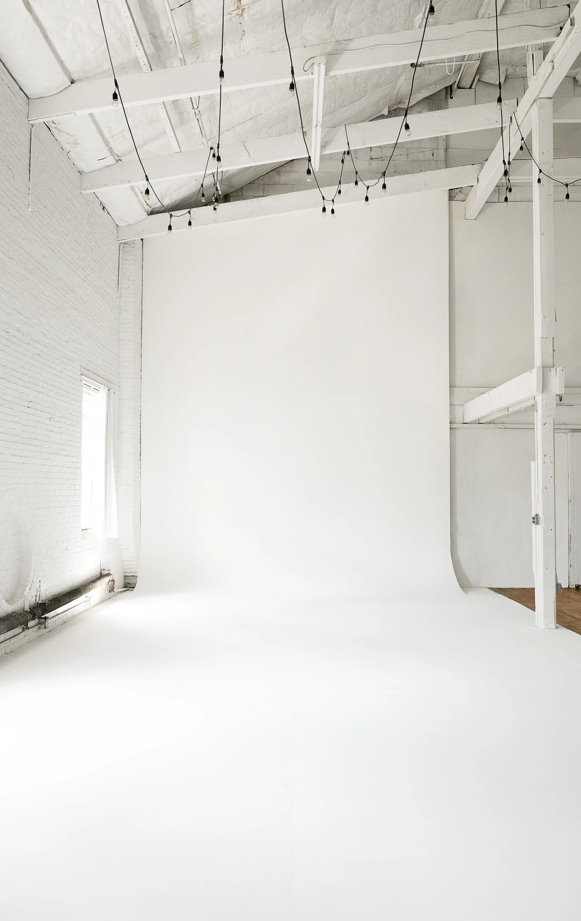 Empty white photo studio with string lights hanging from the ceiling and a white backdrop.