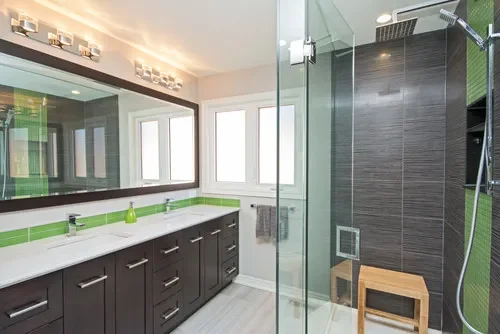 Modern bathroom with a large mirror, double sink vanity with green accents, and a glass-enclosed shower with a wooden stool inside.