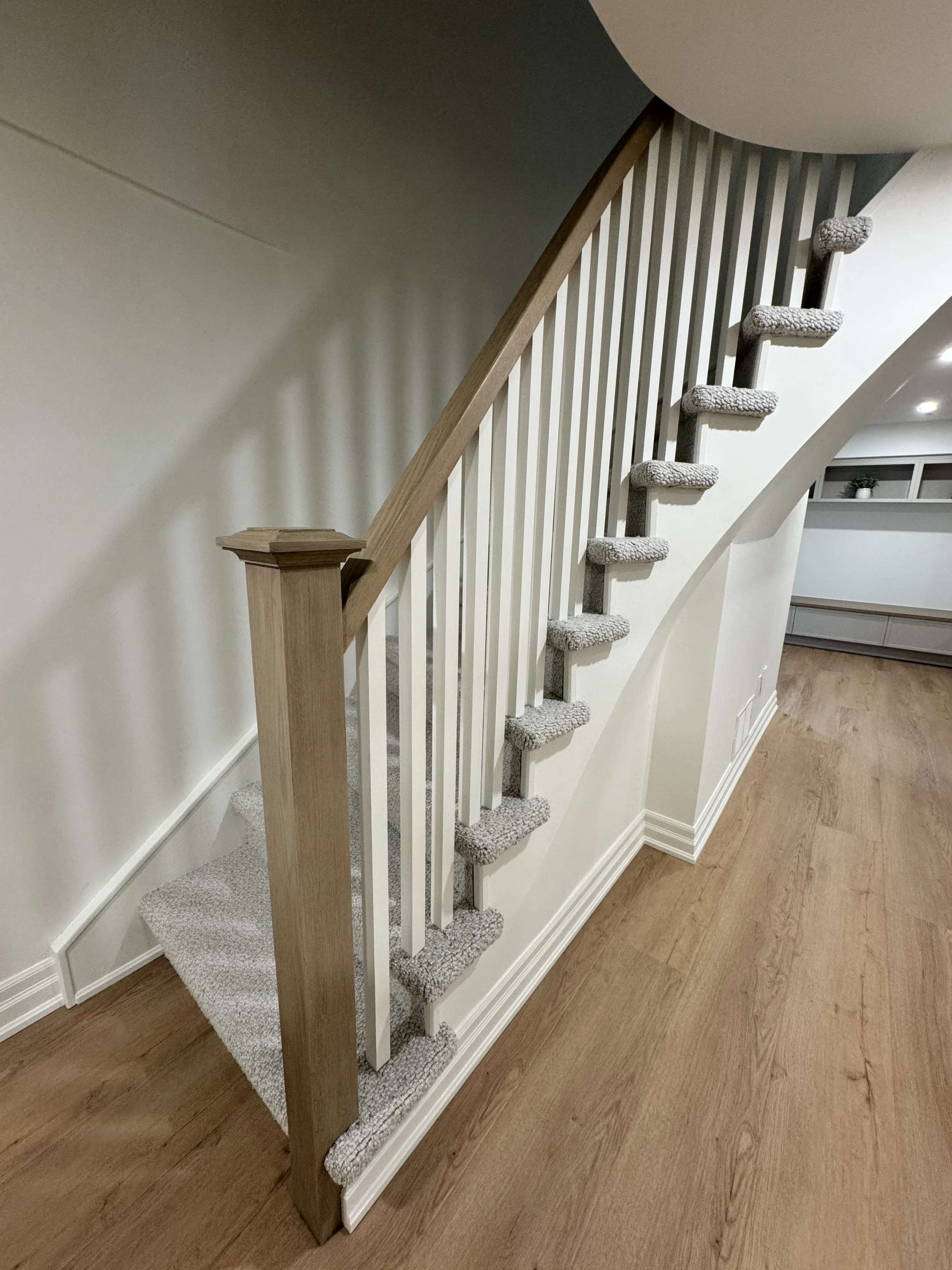 Interior staircase with beige carpeted steps, wooden handrail, white balusters, and hardwood floor nearby.