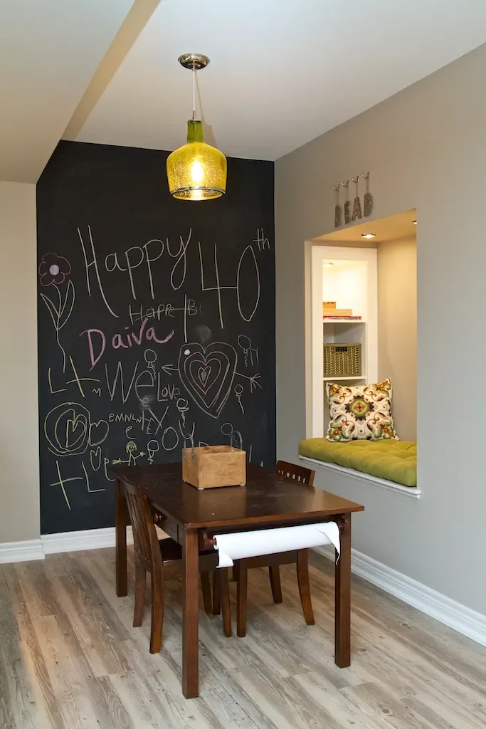 A dining area with a black chalkboard wall that has colorful chalk drawings and birthday message for Daiva's 40th birthday. There's a brown wooden dining table with four chairs, one with a white cushion, and a gold pendant light hanging above. A cozy