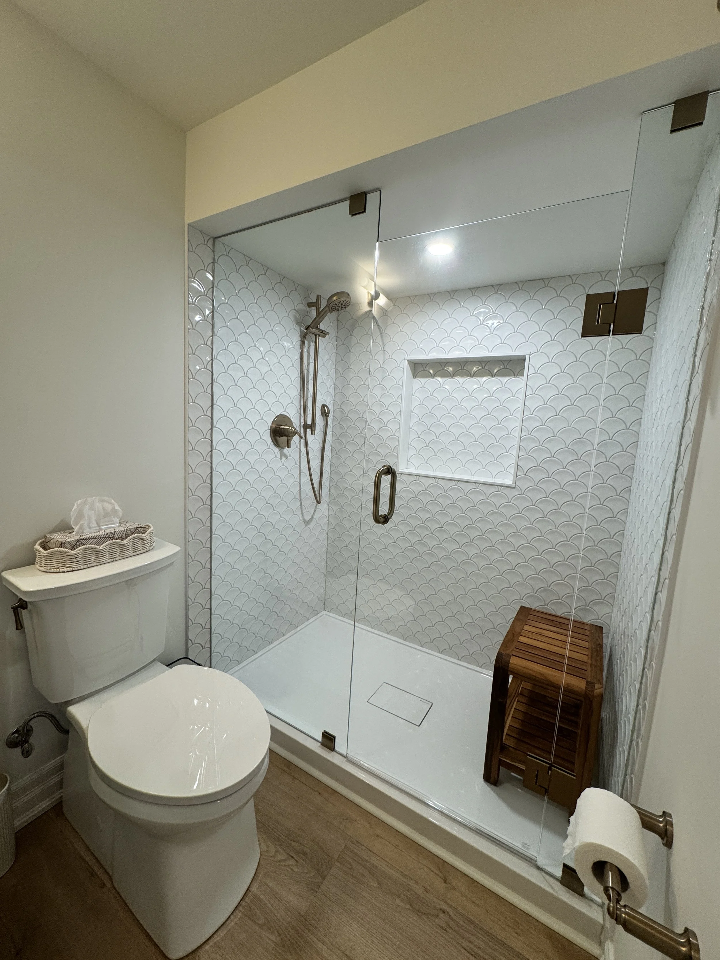 Bathroom with toilet, glass-enclosed shower with a textured white tile wall, wooden stool, tissue box, and toilet paper holder.