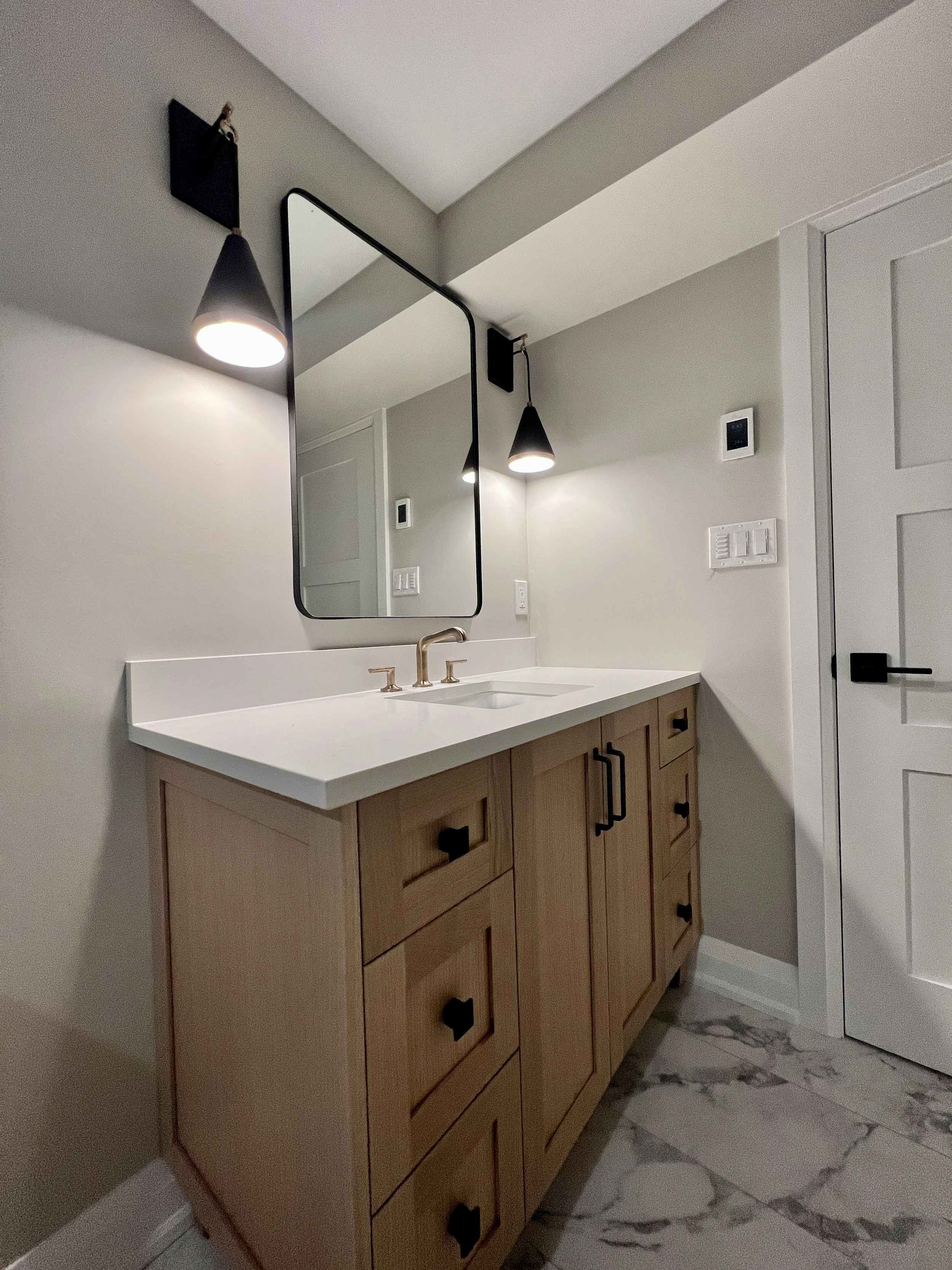 Modern bathroom vanity with a white countertop, a sink with a sleek gold faucet, and wooden cabinets with black handles. Above it is a large rectangular mirror, flanked by black wall-mounted lamps. The wall is painted light gray, and the floor is mar