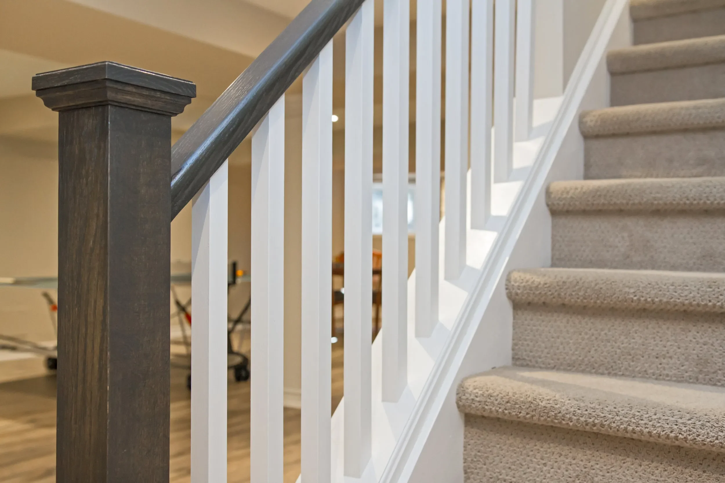 Close-up of a staircase with beige carpeted steps, a dark wooden handrail, and white balusters in an indoor setting.