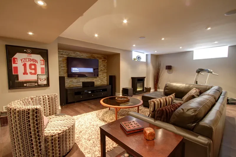 Cozy basement living room with a large leather sofa, patterned armchairs, a flat-screen TV, a fireplace, and exercise equipment, with warm lighting and a framed sports jersey on the wall.