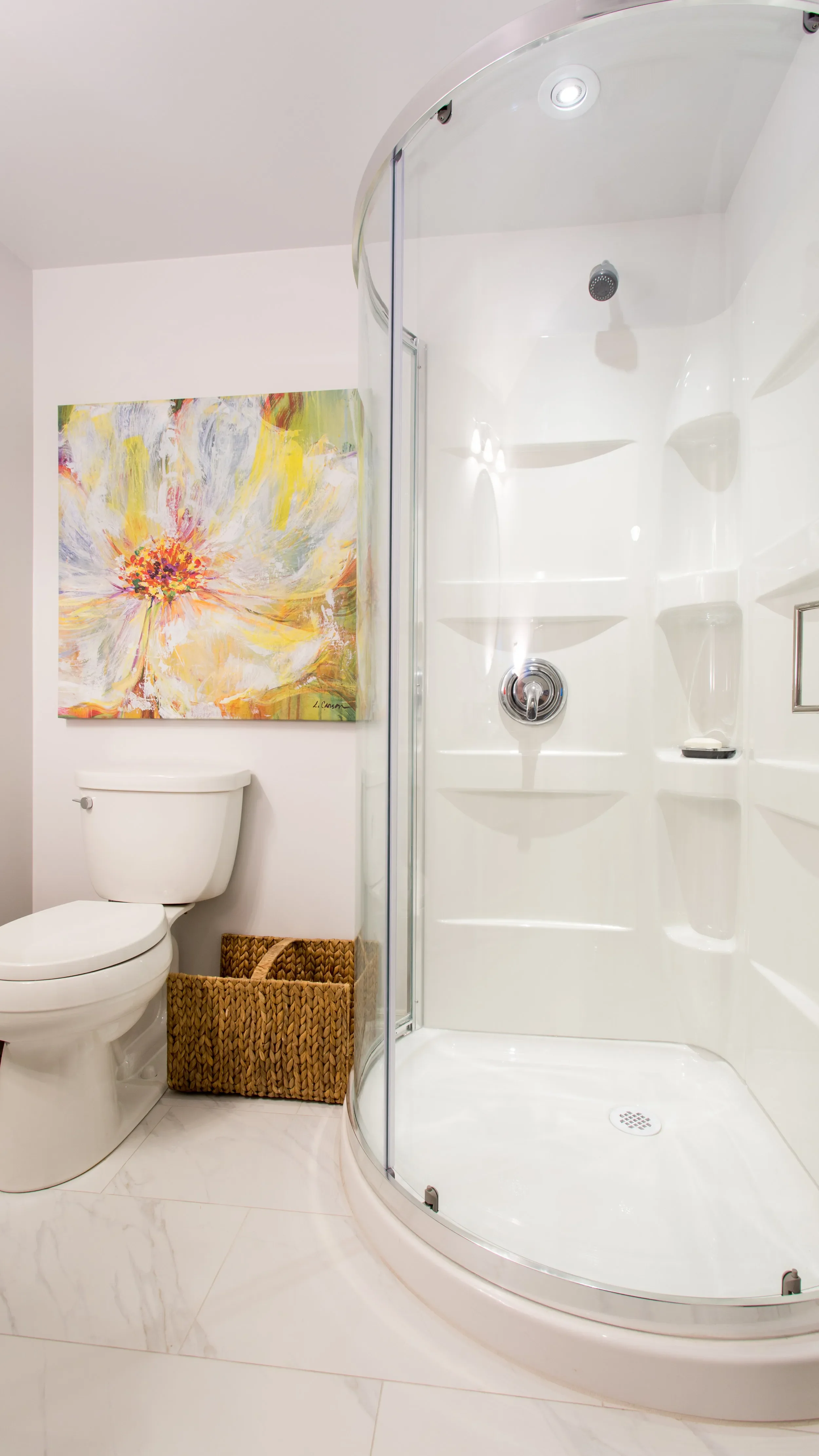 A bathroom with a toilet, a wicker basket, colorful abstract floral wall art, and a corner shower with a curved glass door.