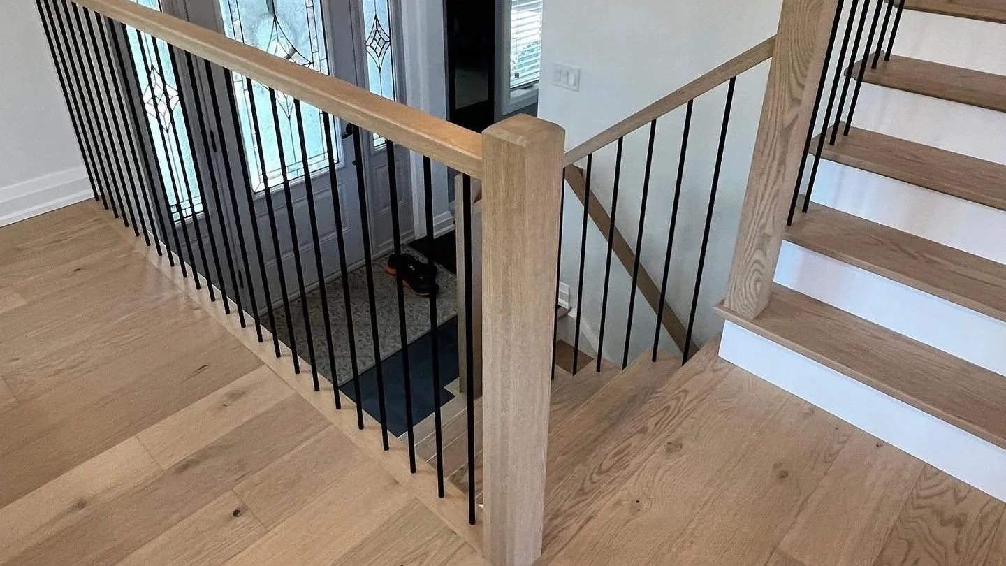 Interior view of a staircase with wooden steps and black metal railing, looking down from the top floor, showing a front door with decorative glass panels at the landing.