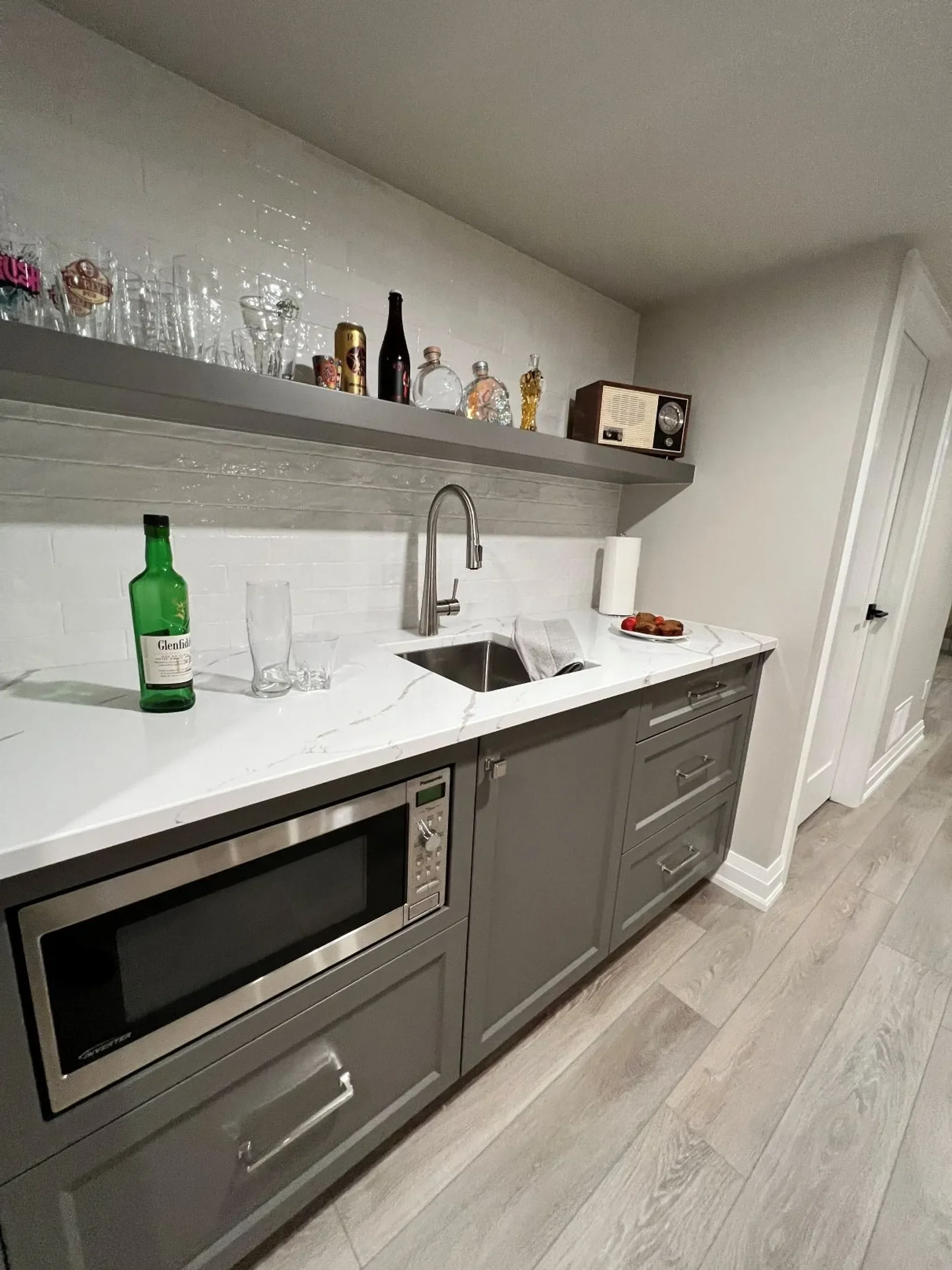 A modern kitchen with gray cabinets, a white marble countertop, a sink with a faucet, a microwave, a bottle of Glenfiddich whiskey, and a paper towel roll. There are glasses and bottles on a gray open shelf above the countertop. A plate of food is on