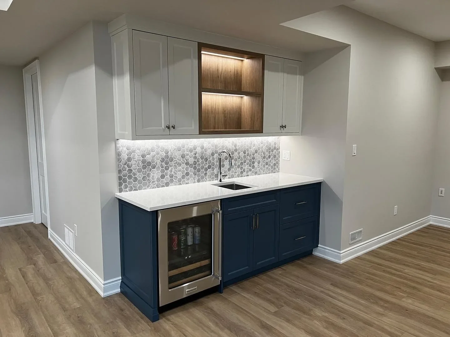 Small kitchenette with gray and blue cabinets, white countertop, hexagonal tile backsplash, under cabinet lighting, and wine cooler, in a room with wood flooring and white walls.