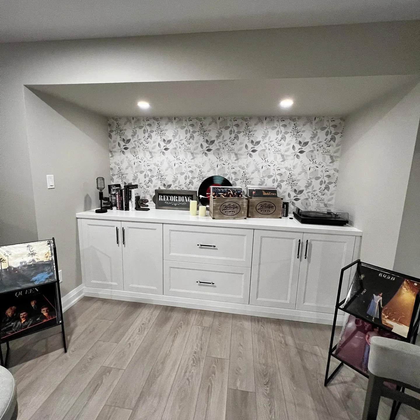 A white console table in a room with overhead lighting, decorated with vinyl records, books, candles, and a record player, against a wallpapered wall with leaf patterns.