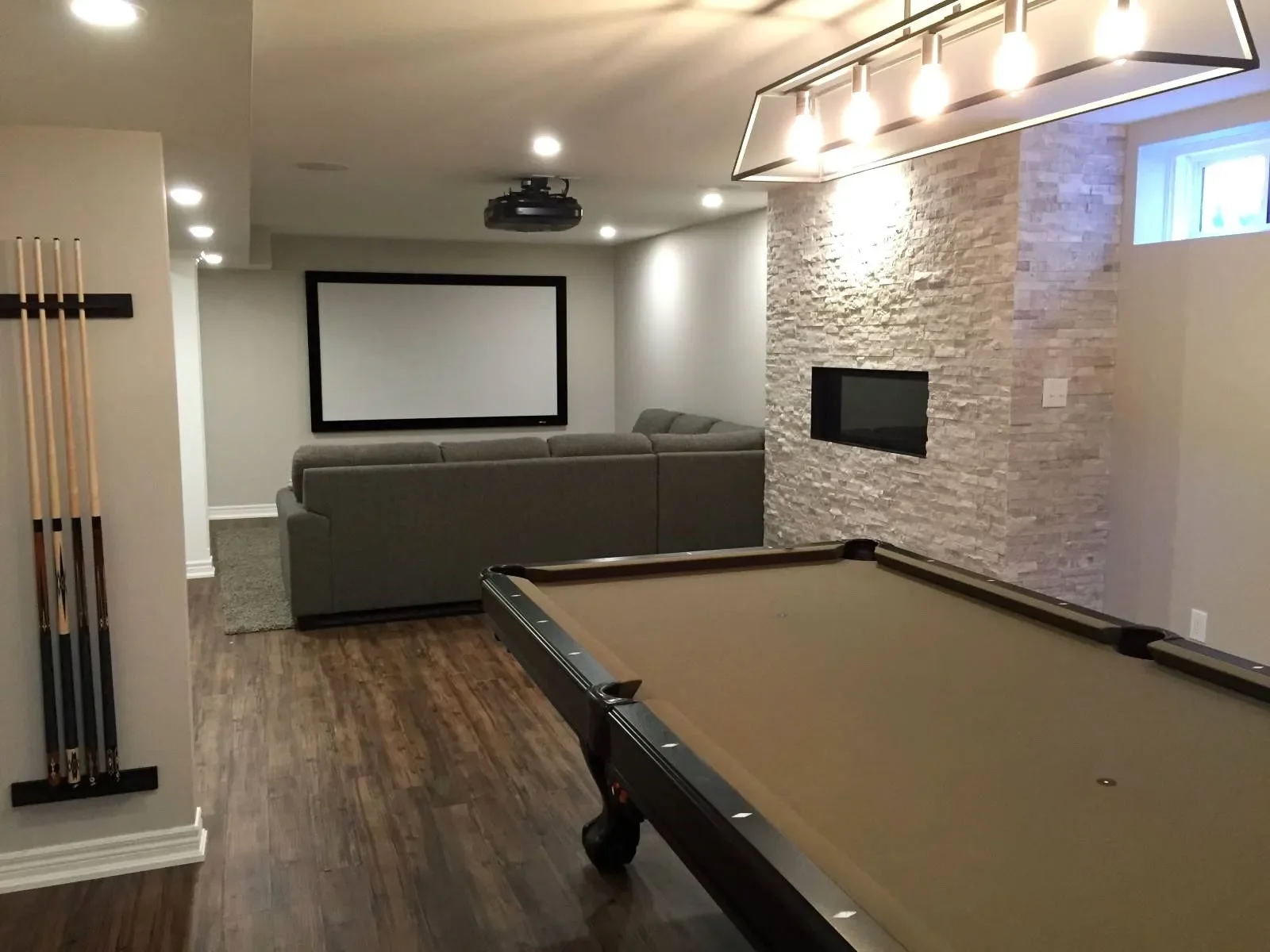 A basement entertainment room with a pool table in the foreground, a gray sectional sofa facing a large projector screen on a white wall, a mounted fireplace on a textured stone wall, and recessed lighting overhead.