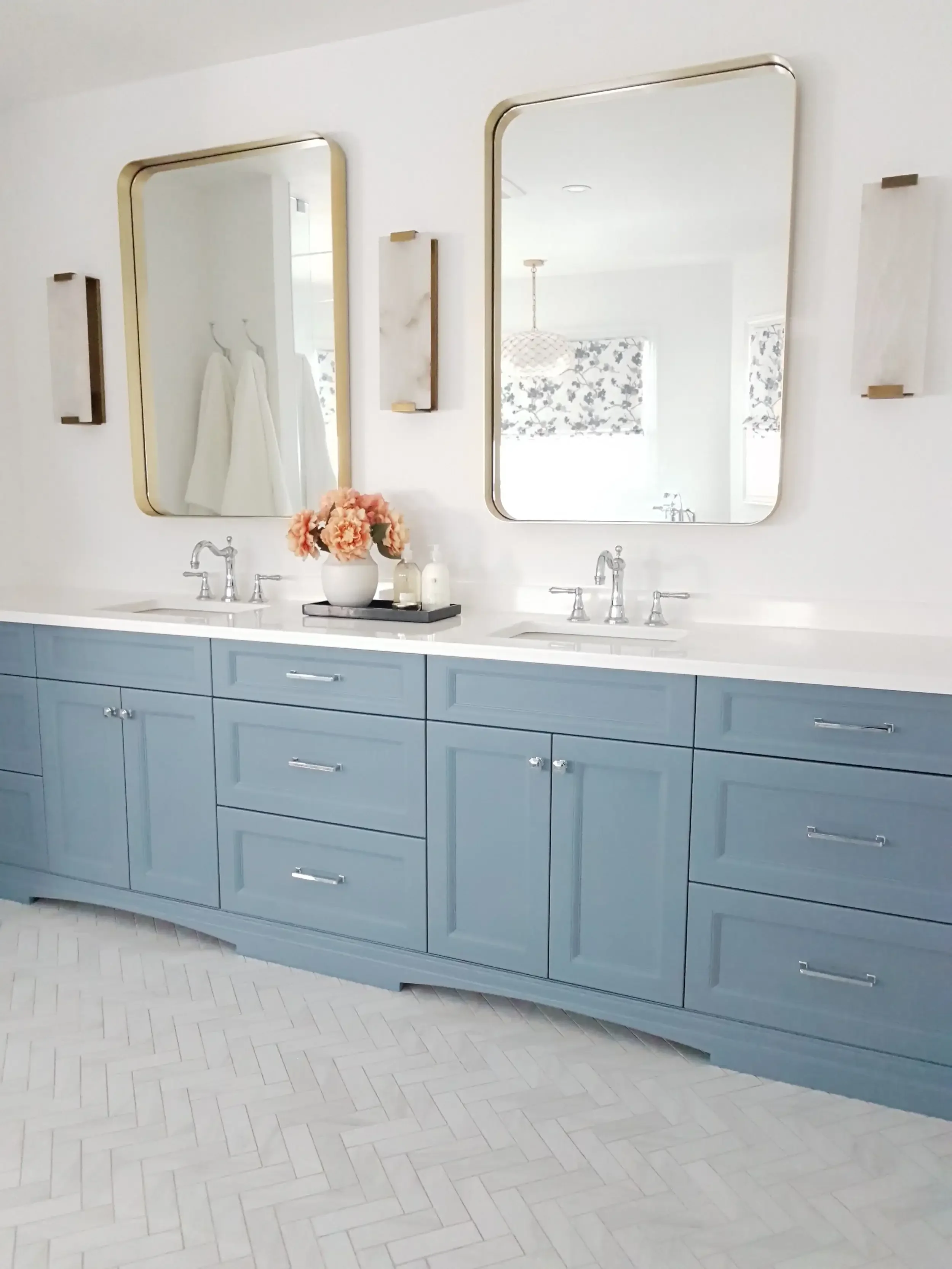 Light blue bathroom vanity with dual sinks, silver faucets, and eight drawers, topped with a white countertop. Two large rectangular mirrors with gold trim hang above the sinks, flanked by decorative wall sconces. A white vase with pink flowers and lotion bottles sit on a tray on the countertop. The floor is white tiled in a herringbone pattern.