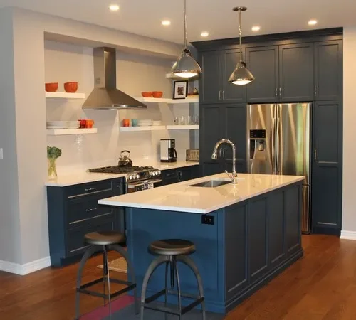 Modern kitchen with navy blue cabinets, white countertops, and stainless steel appliances, including a refrigerator and range hood. Open shelves with colorful dishes, two pendant lights, and a kitchen island with two stools.