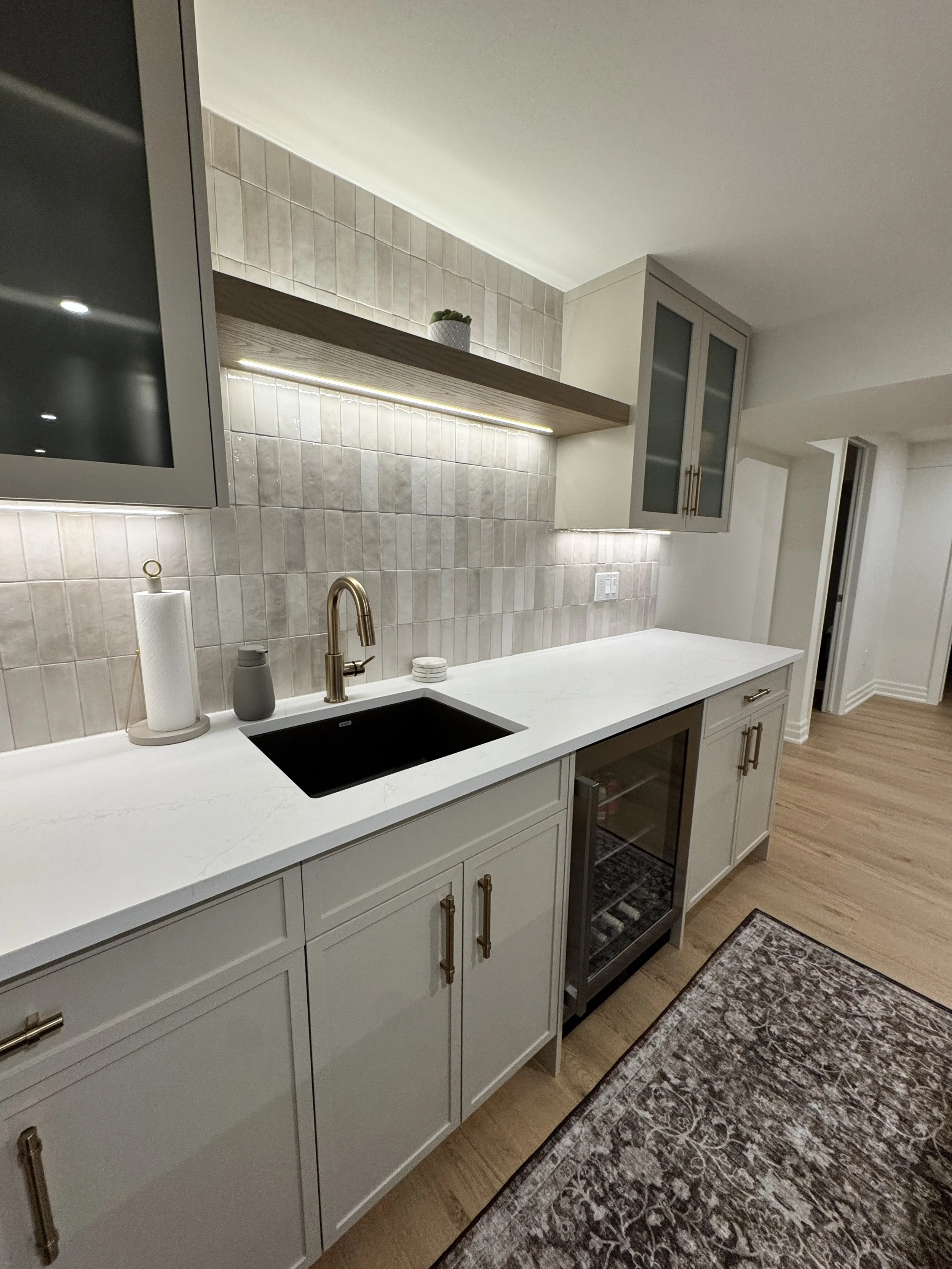 Modern kitchen with white cabinets, black sink, gold faucet, beige tiled backsplash, and small potted plant on a wooden shelf.