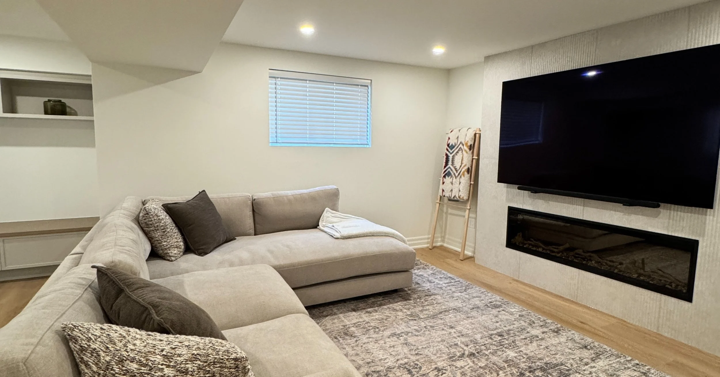 A cozy living room with a beige sectional sofa decorated with various throw pillows, a window with blinds, a ladder with a patterned blanket, a large flat-screen TV mounted on a textured wall, a fireplace below the TV, and a patterned rug on wooden f
