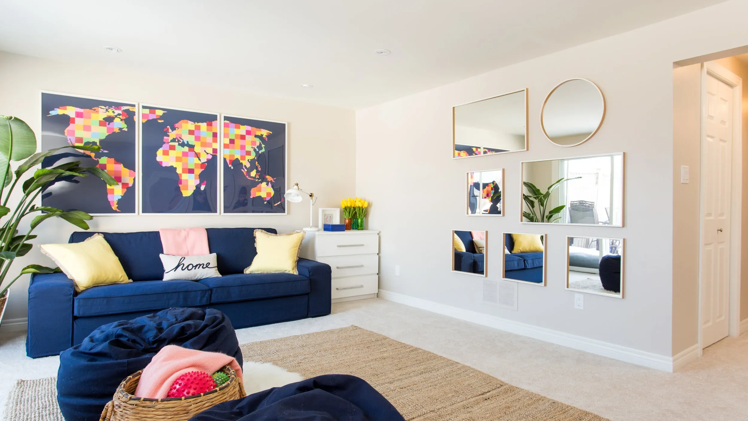 Living room with a dark blue sofa, yellow and pink pillows, and wall art of a colorful world map. There are mirrors on the wall, a white dresser with yellow tulips, and a large indoor plant. The room has a beige carpet and white walls.