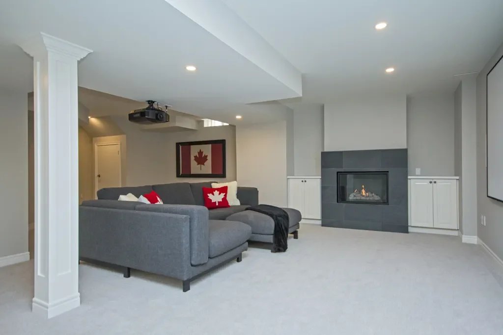 Living room with gray sectional sofa, Canadian flag-themed pillows, fireplace, white built-in cabinets, projection system, and framed Canadian flag on the wall.