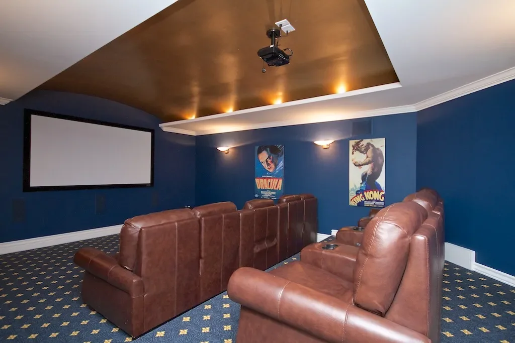 Home theater with brown leather recliners facing a large white projection screen on a blue wall with vintage movie posters, carpeted floors, and a ceiling-mounted projector.