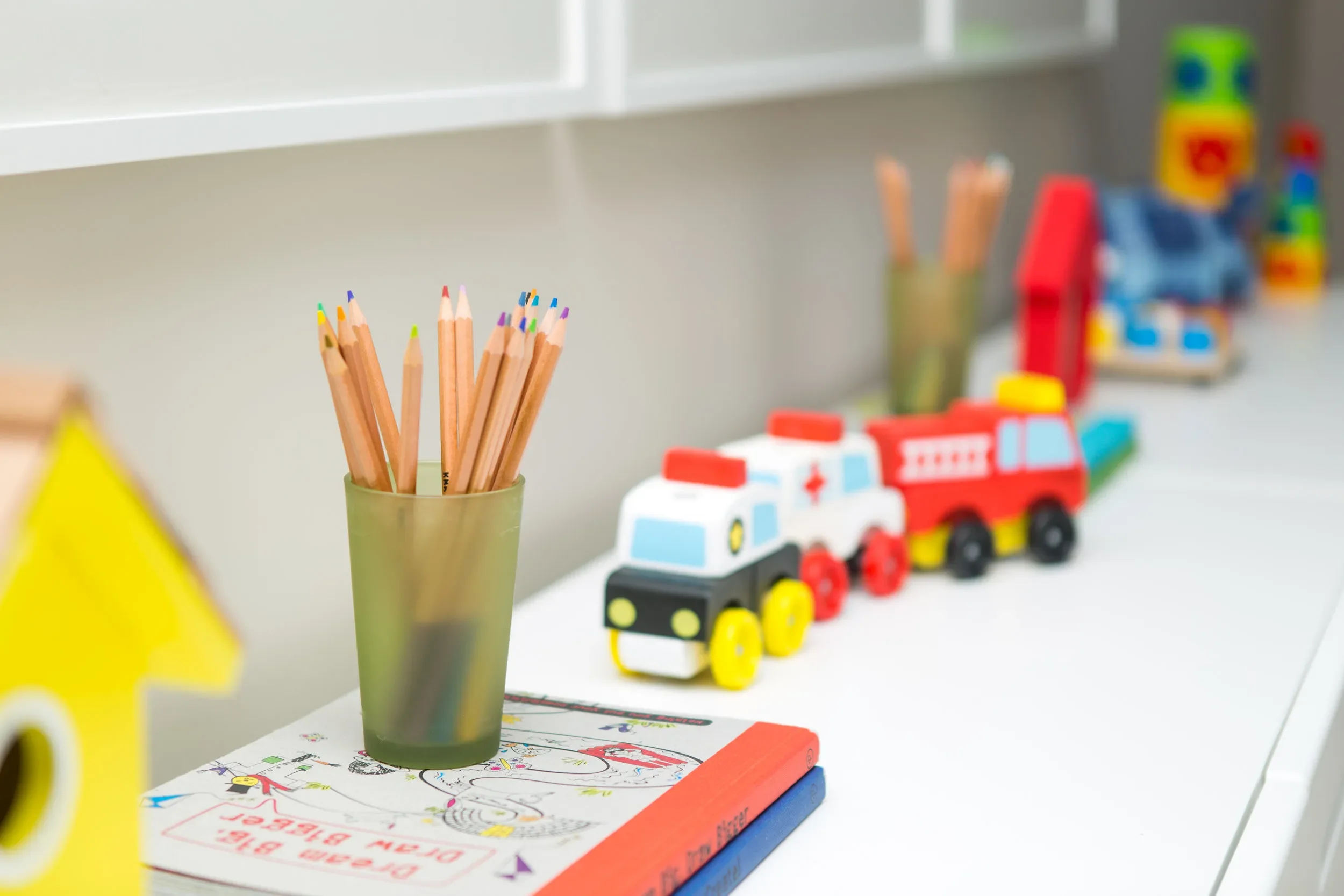 Colorful pencils in a green cup, a children's drawing on the table, and toy emergency vehicles including a fire truck and ambulance, with blurred colorful building blocks in the background.