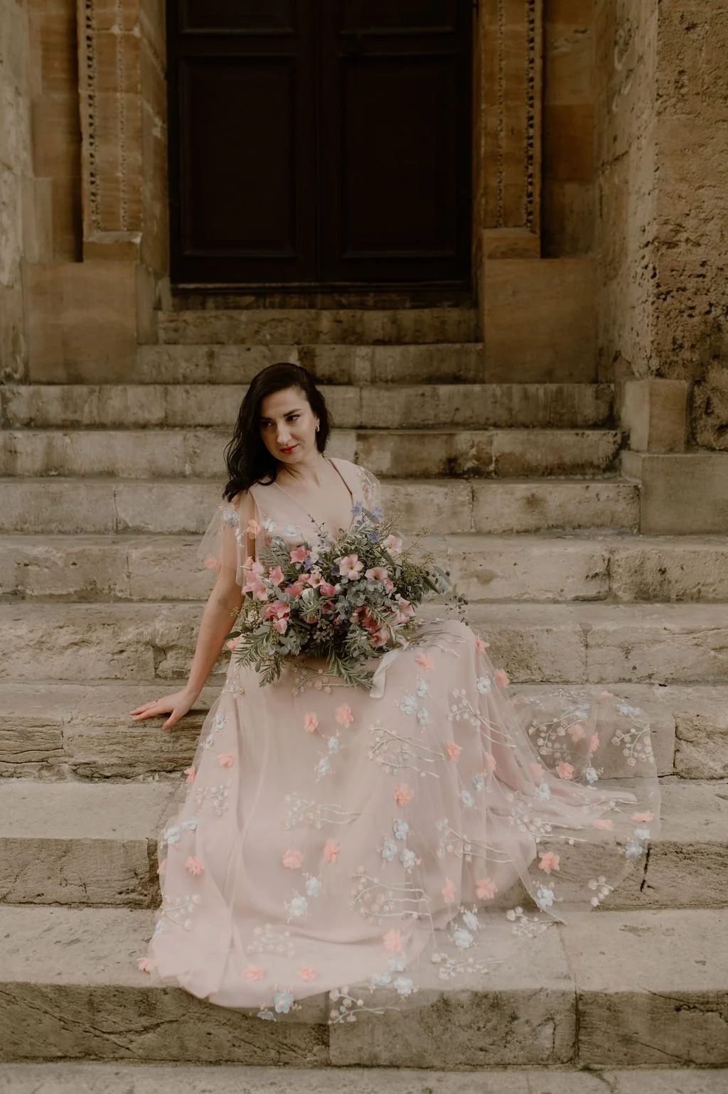 A woman in a pink floral dress sitting on stone steps holding a bouquet of flowers.
