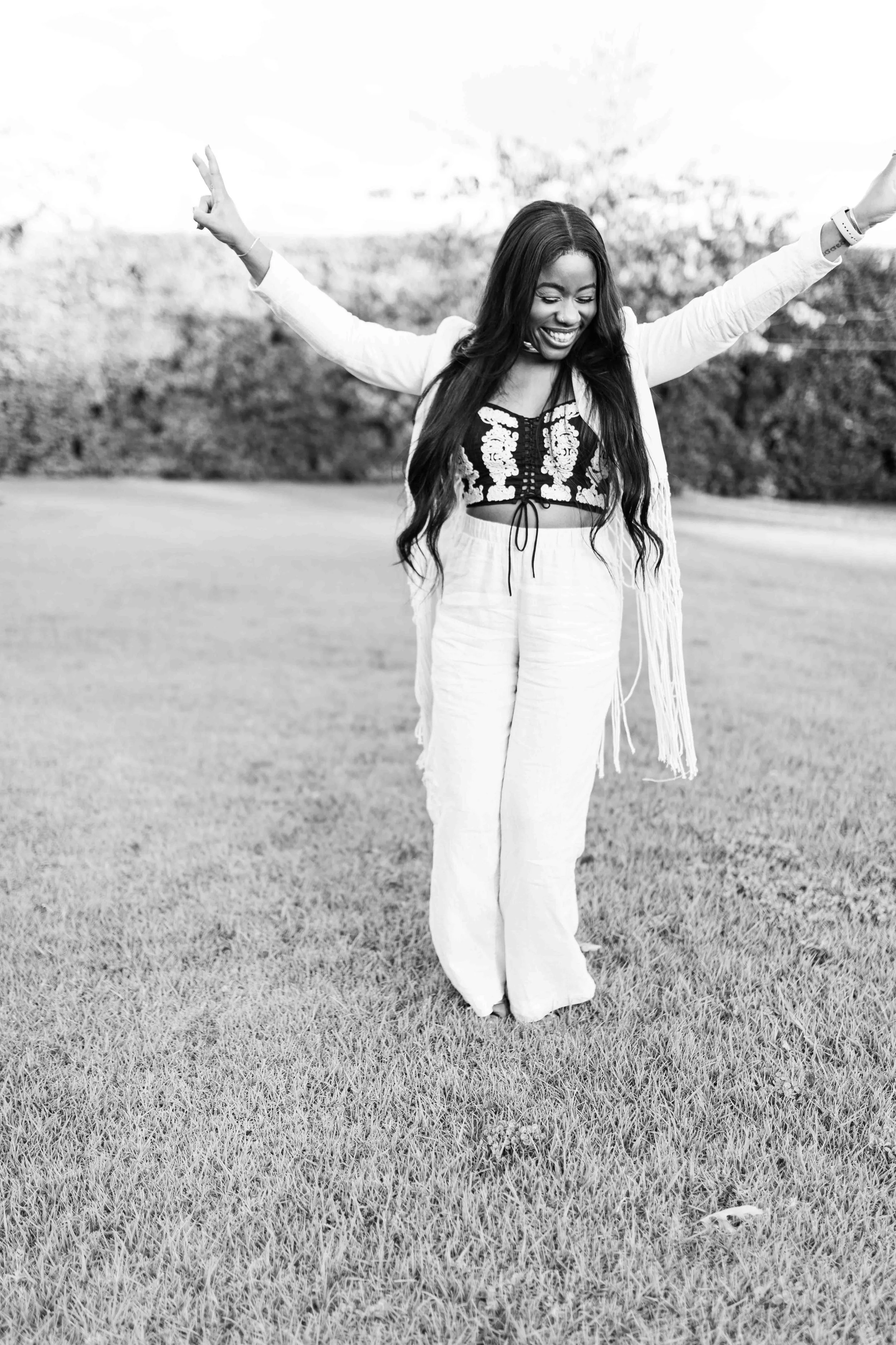 A black and white photograph taken during a baby shower event showing a woman with long hair smiling and standing on grass with her arms raised.