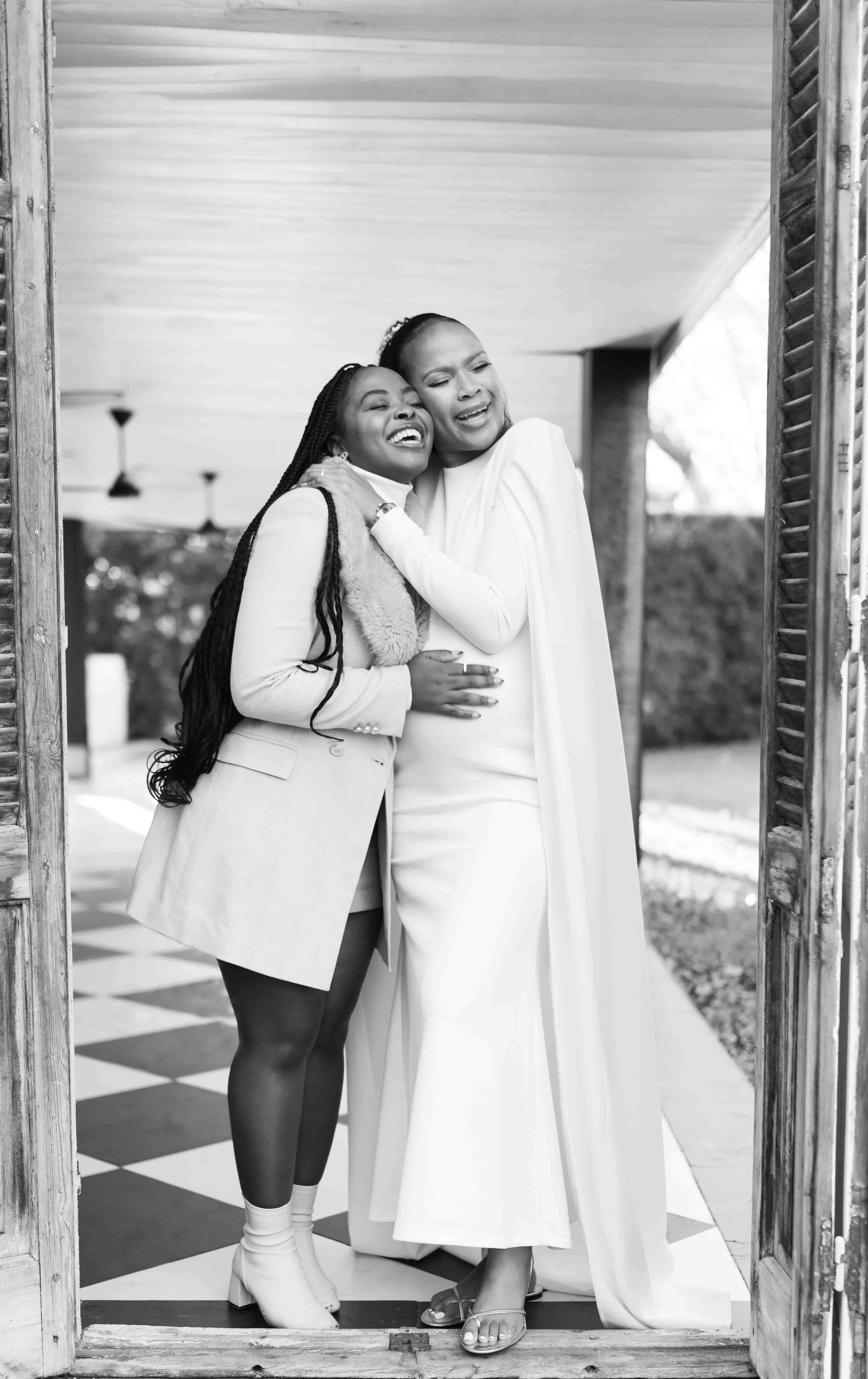 A black and white photograph taken during a baby shower event showing two women hugging and smiling in a doorway, with a porch and garden in the background.