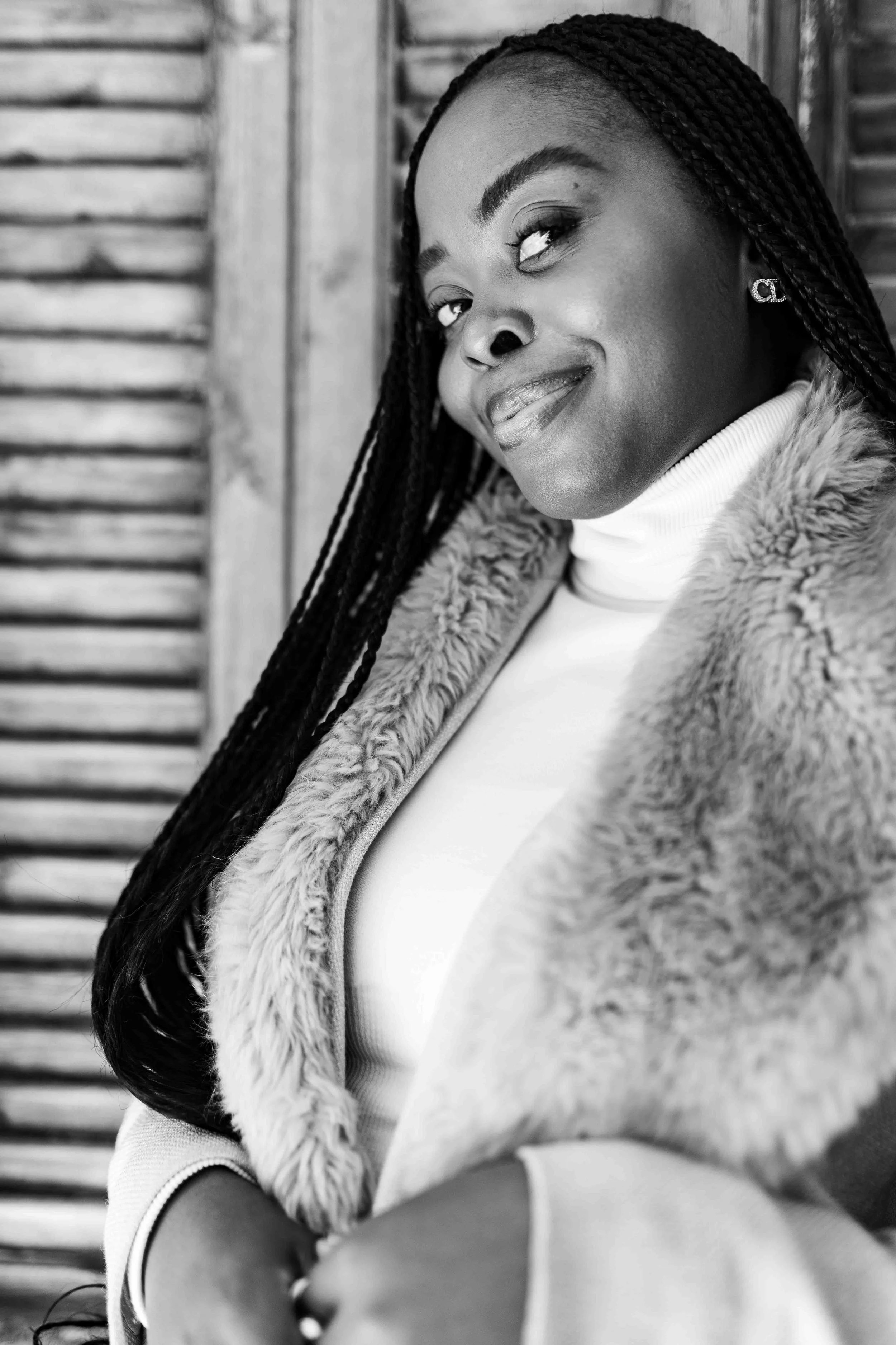 A black and white portrait photograph taken during a baby shower event showing a woman with long braided hair, wearing a fur coat and a turtleneck, smiling slightly with a confident expression, standing in front of a wooden background.