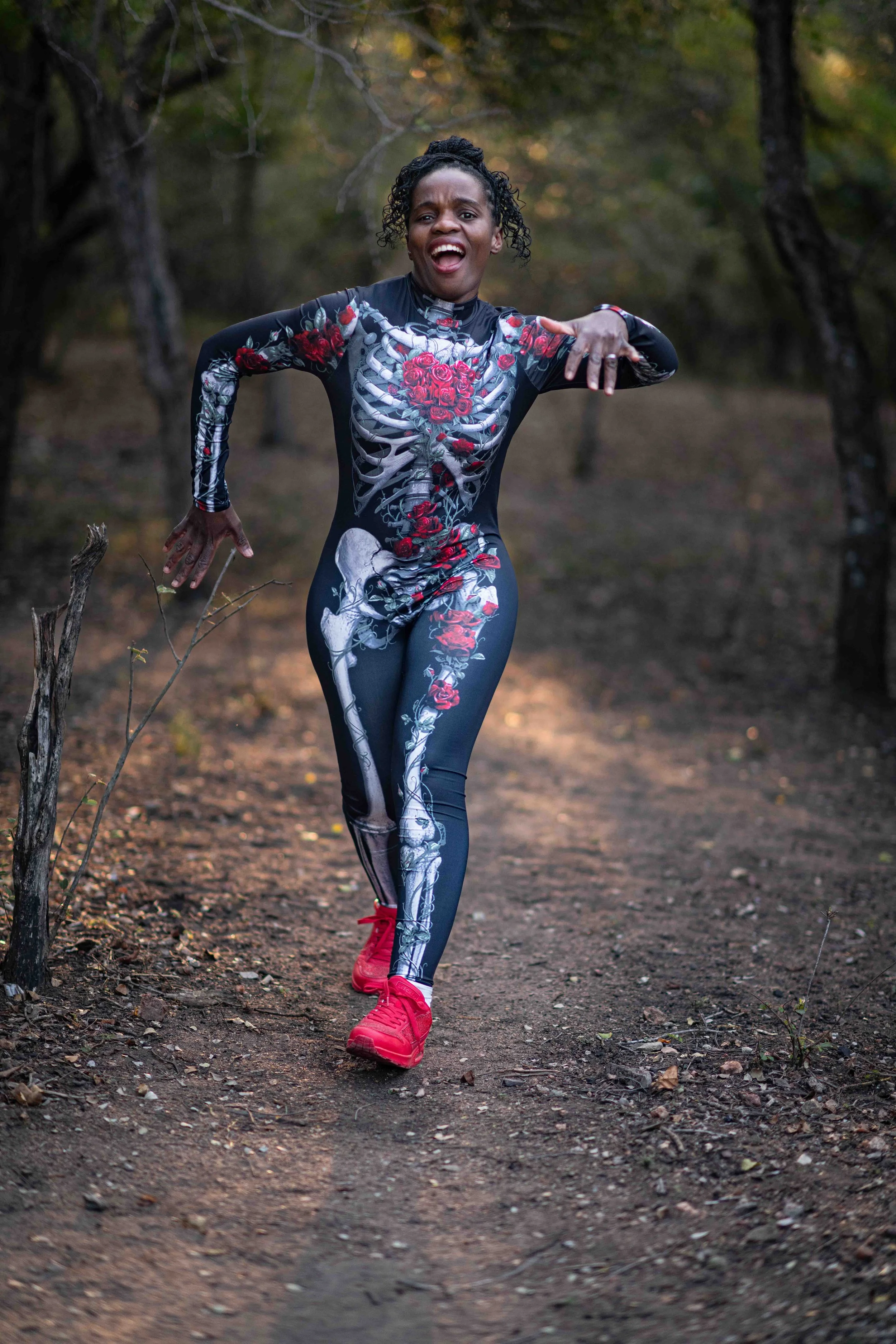A photoshoot of a women in costumes at a birthday party, making playful a pose outdoors at the Kruger National Park, wearing a bodysuit with a skeleton and roses design, and red sneakers
