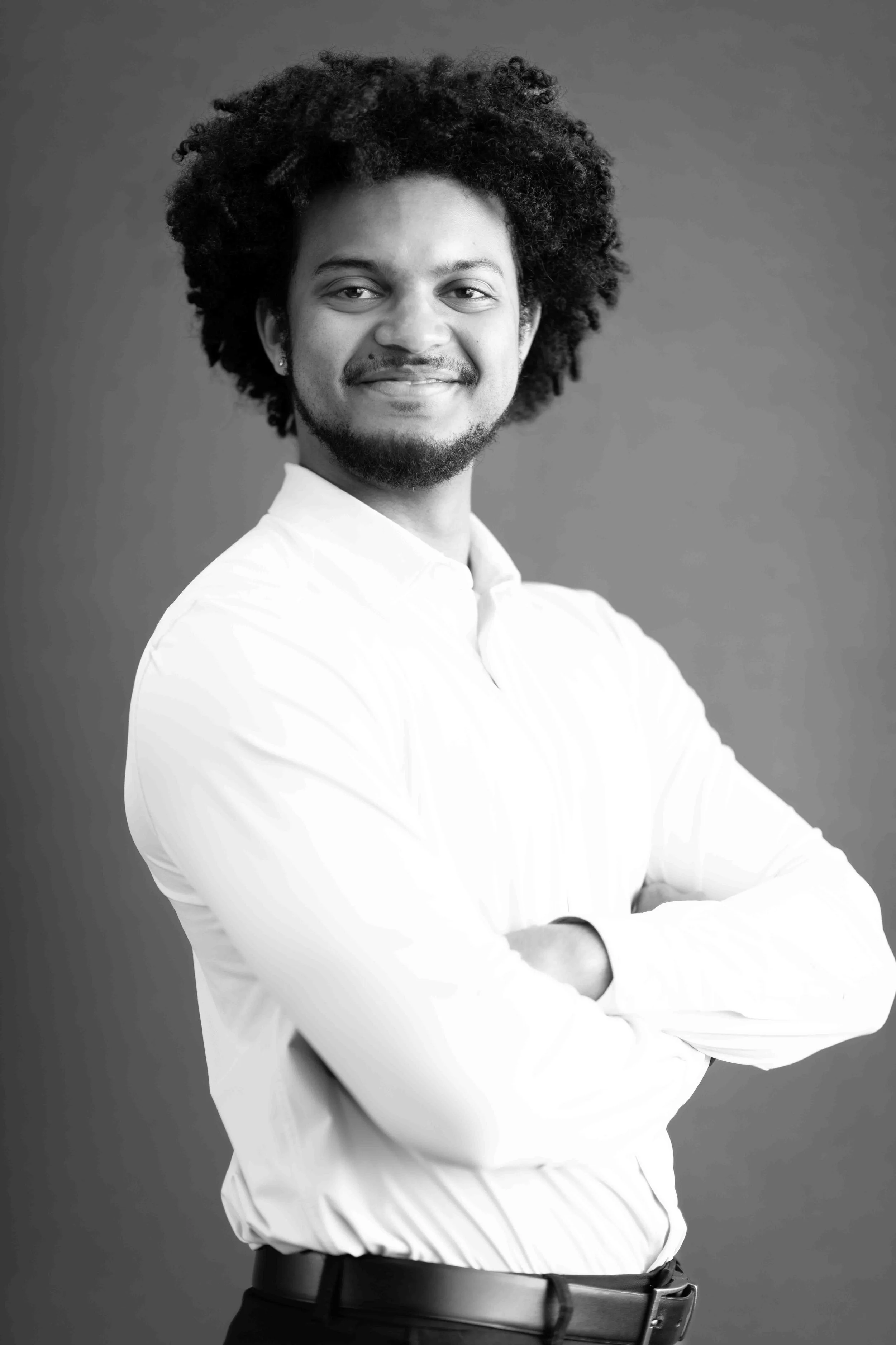 A corporate professional headshot taken in Johannesburg in black and white of a young man with curly hair, smiling, wearing a white shirt with arms crossed, standing against a plain background.