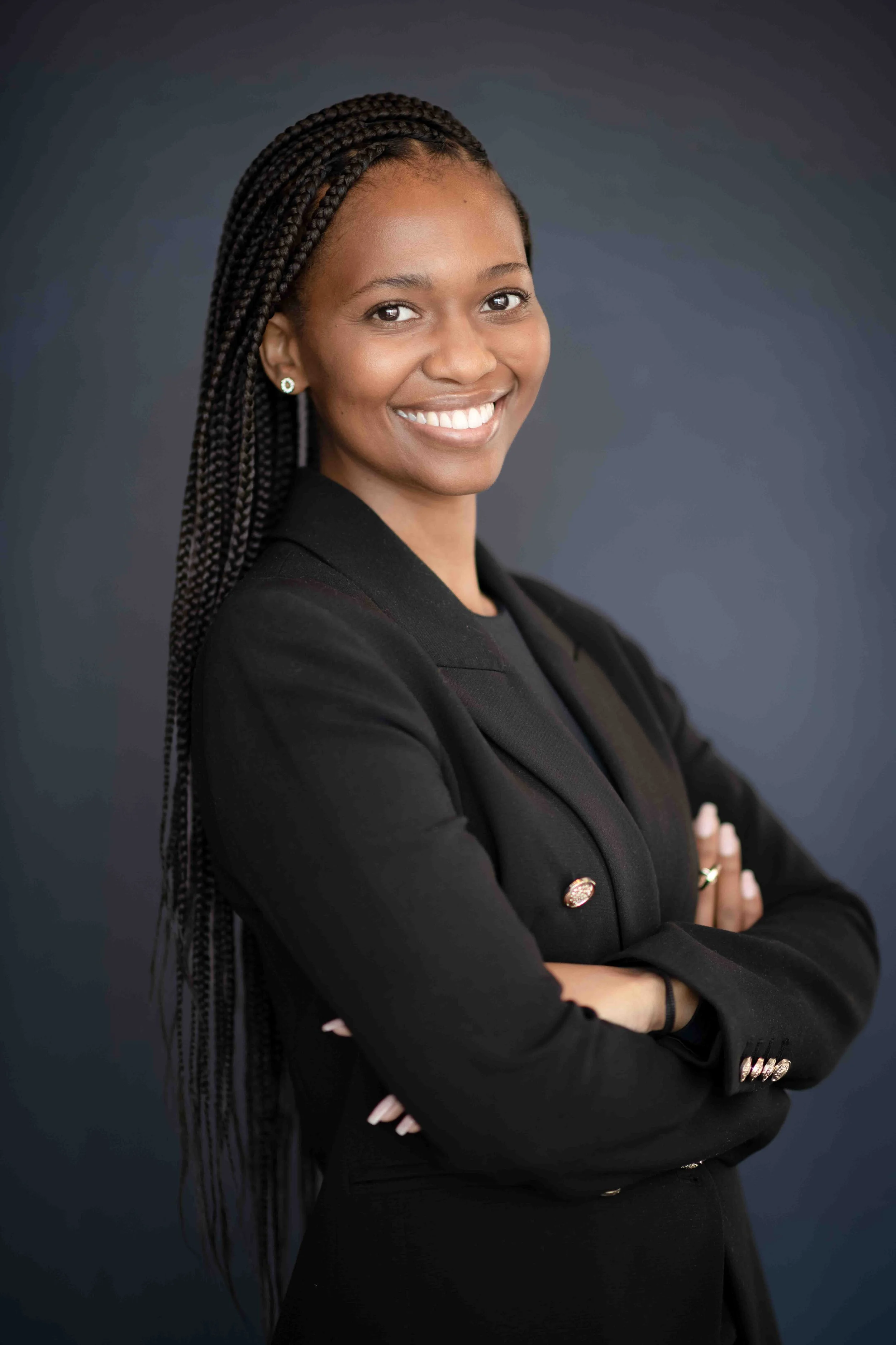 A corporate professional headshot taken in Cape Town in black and white of a woman with braided hair, wearing a black blazer, smiling, with her arms crossed against a dark background.
