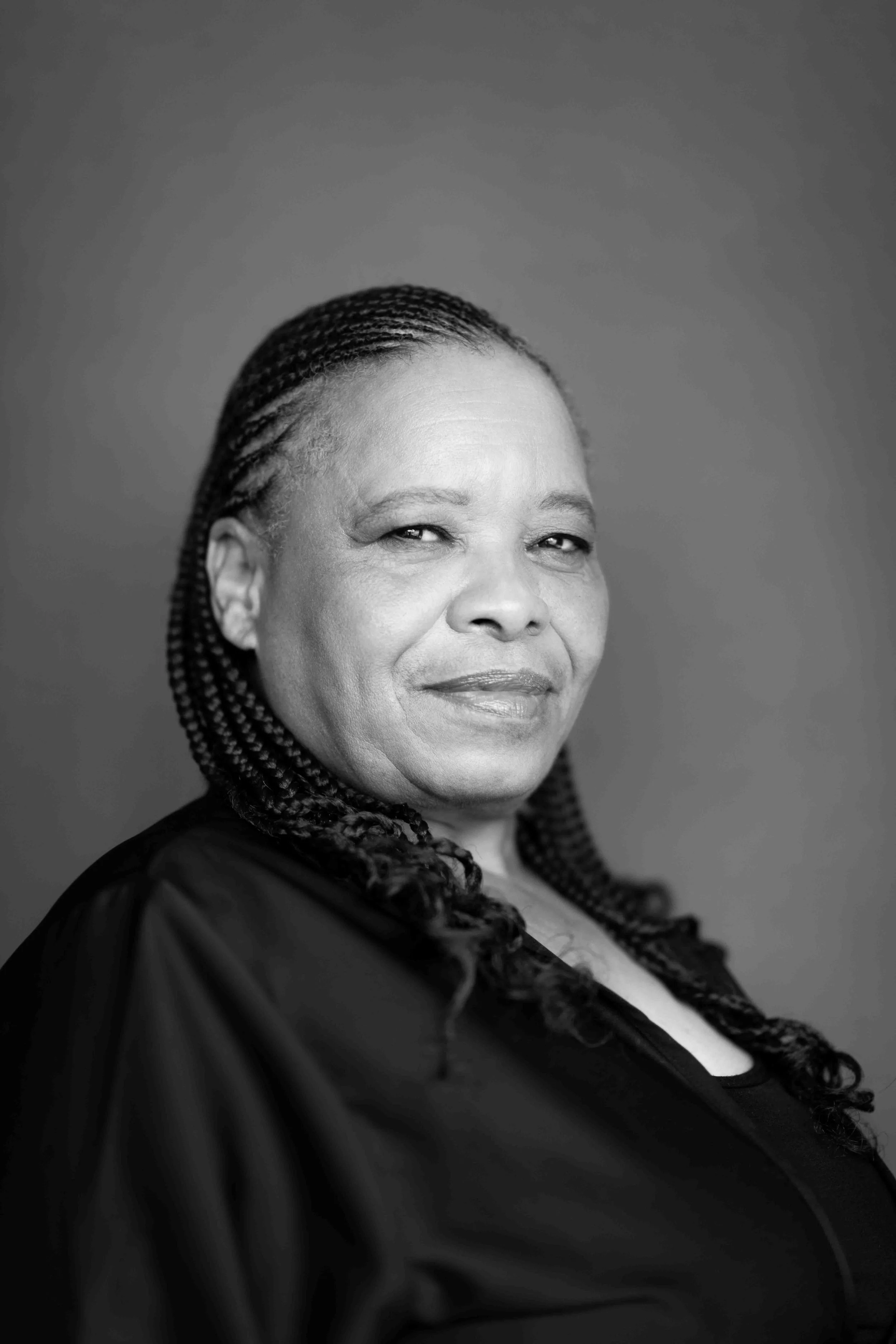 A corporate professional headshot taken in Cape Town in black and white of a confident woman with braided hair, wearing a dark top, against a plain background.