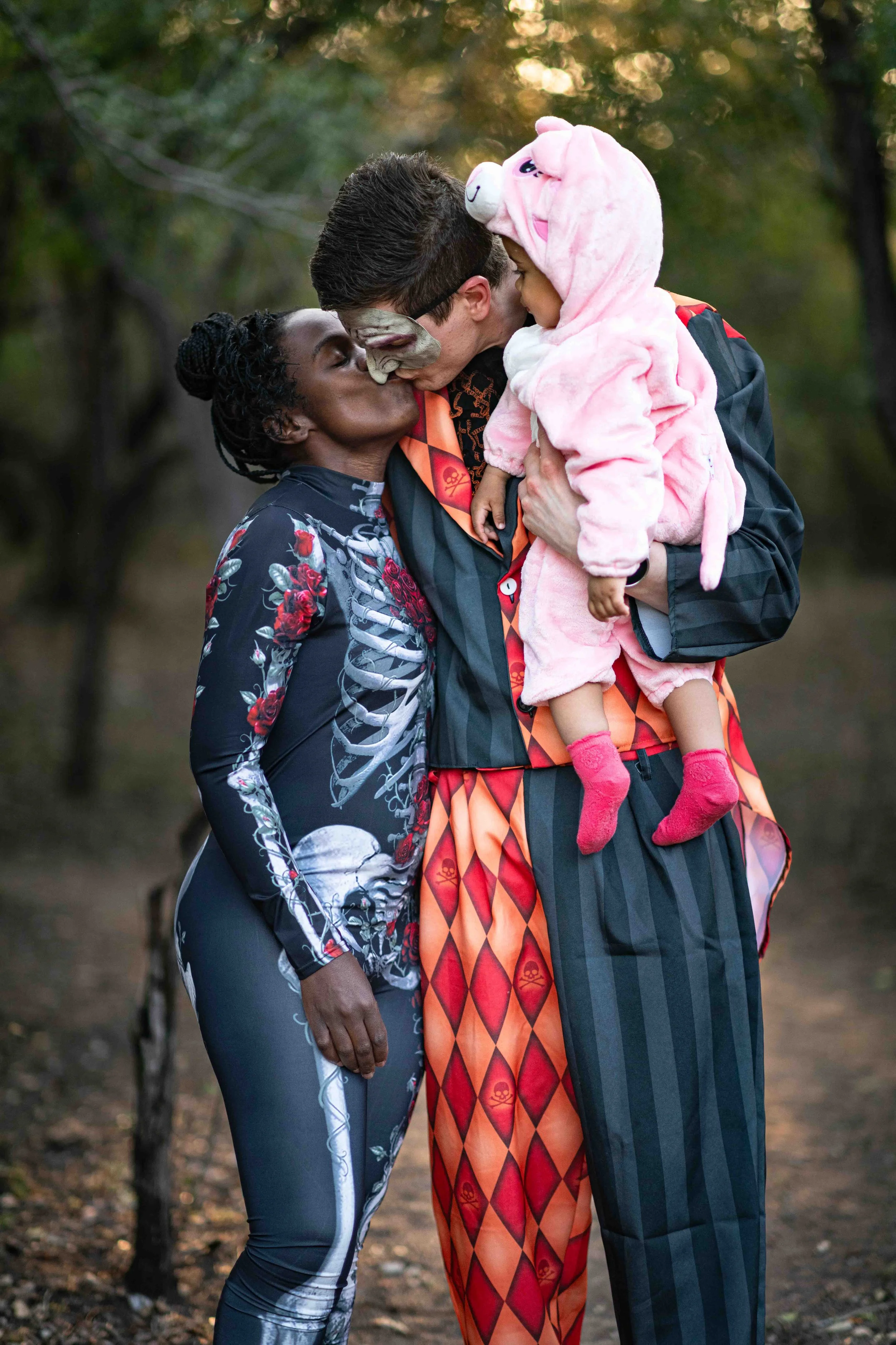 A photoshoot of a family in costumes at a birthday party, outdoors at the Kruger National park. The couple kiss, while holding their daughter.