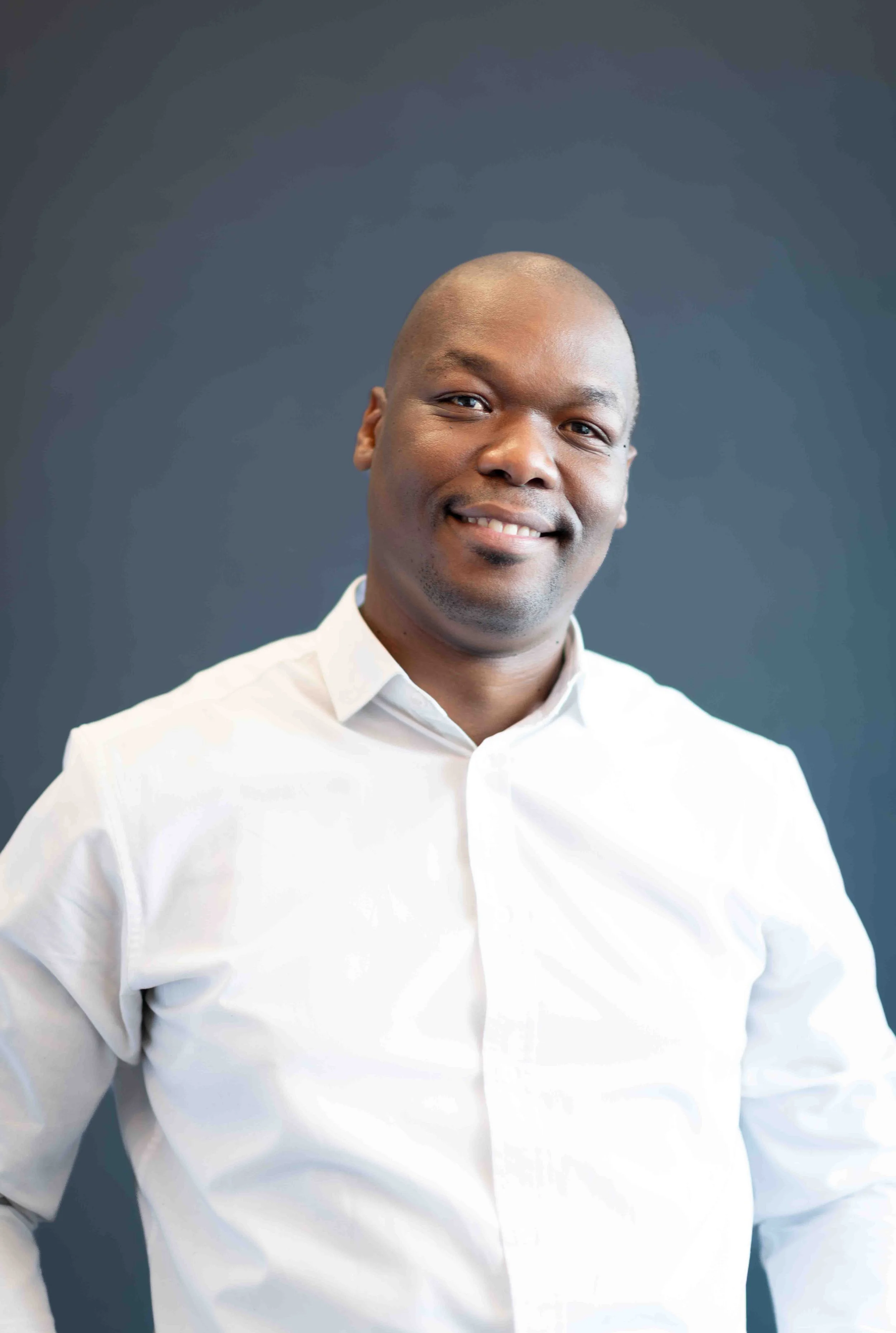 A corporate professional headshot taken in Johannesburg of a smiling man wearing a white button-up shirt, standing against a dark background.
