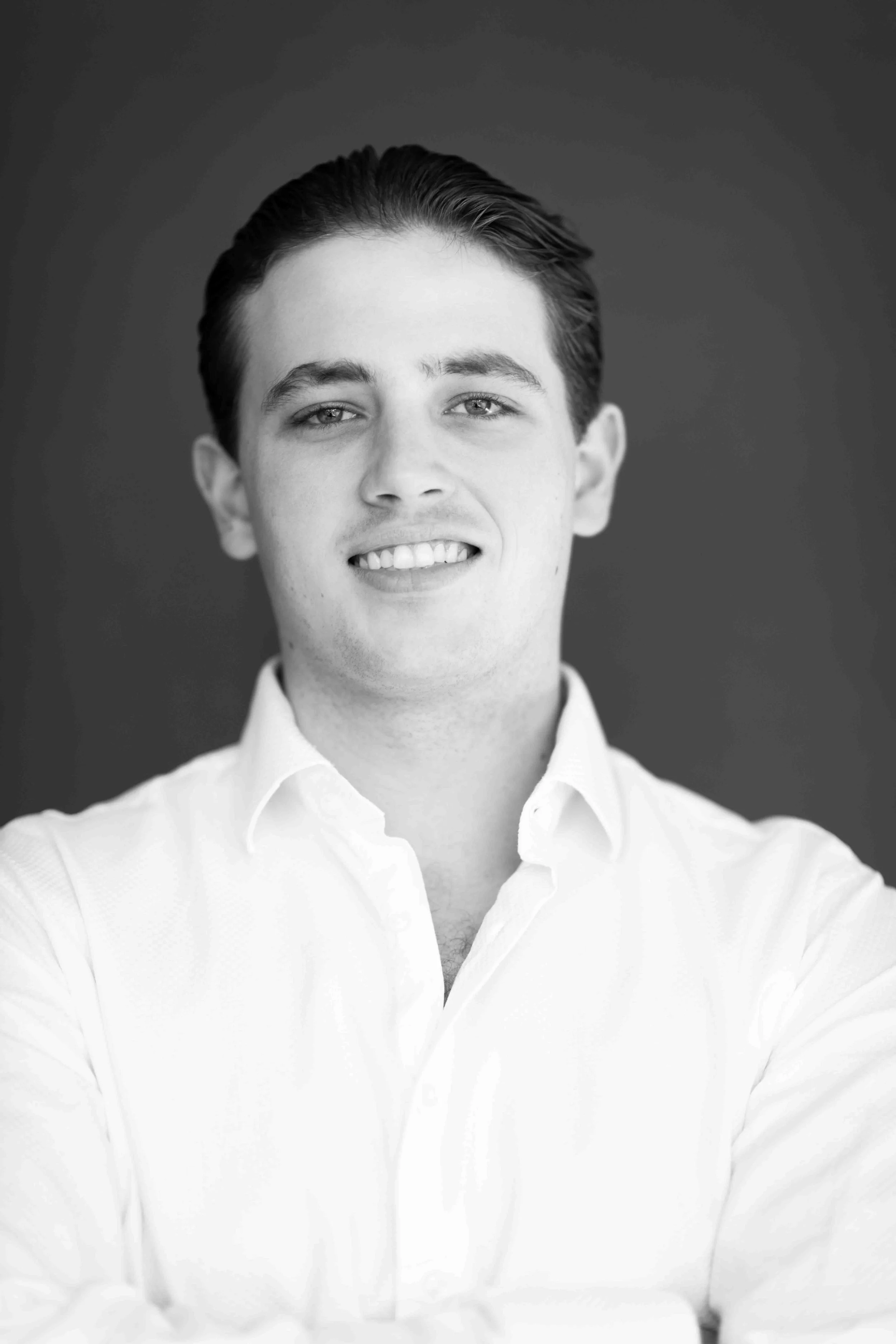 A corporate professional headshot taken in Cape Town in black and white of a young man with slicked back hair, wearing a white button-up shirt, smiling slightly, against a dark background.