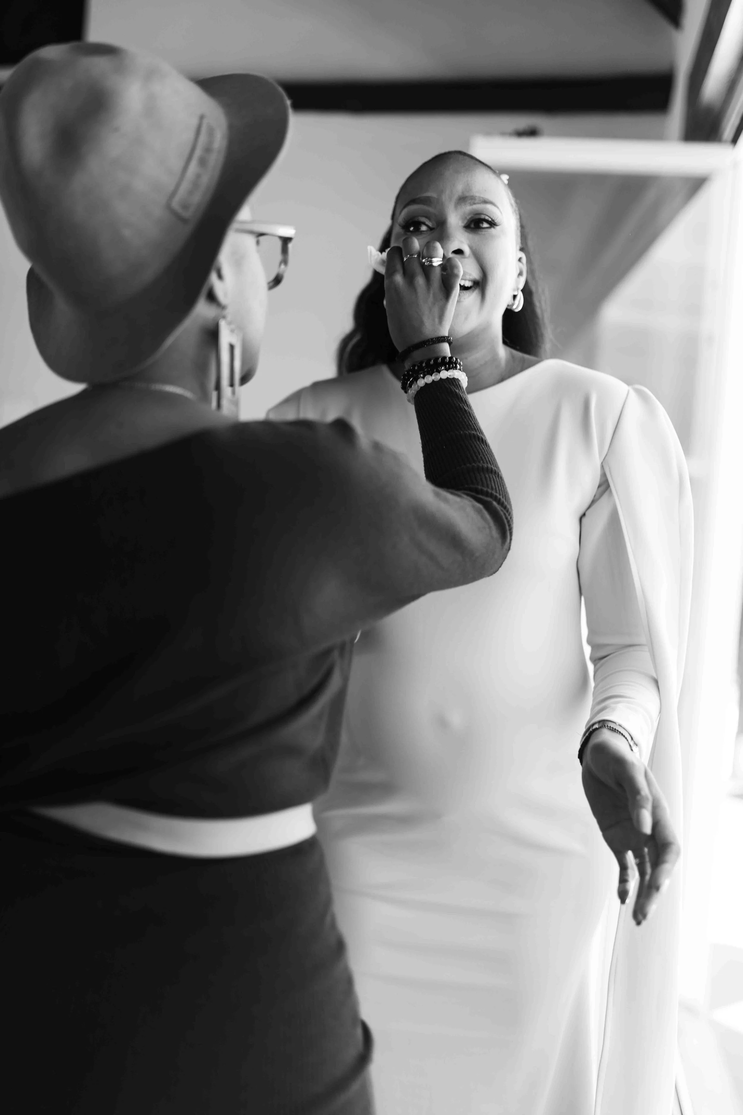 A photograph taken during a baby shower event showing s woman in a white dress having her tears dried off by her sister in law in a room with natural light.