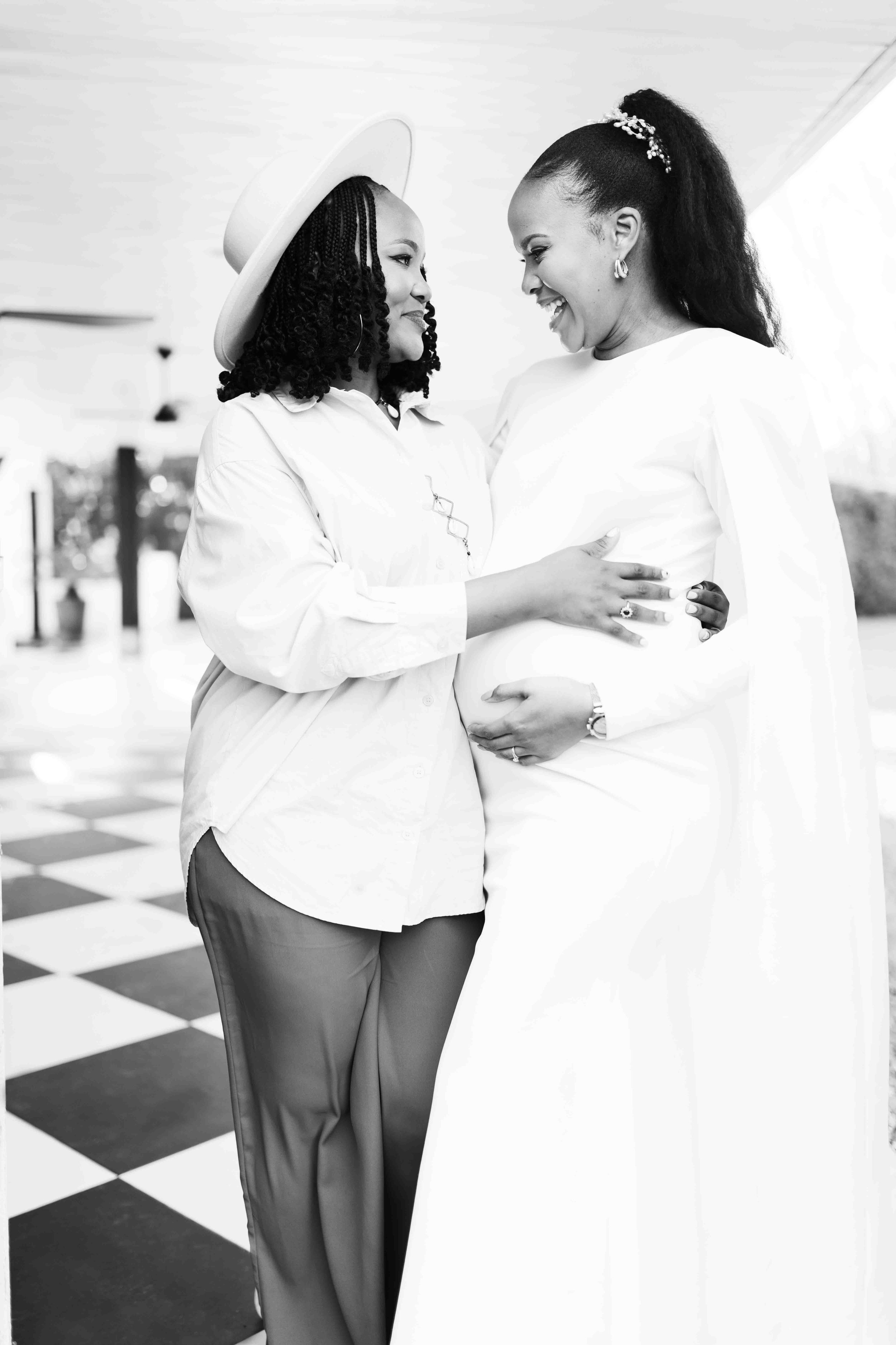 A black and white photograph taken during a baby shower event showing two women, one pregnant, sharing a special moment indoors, smiling and touching her belly.