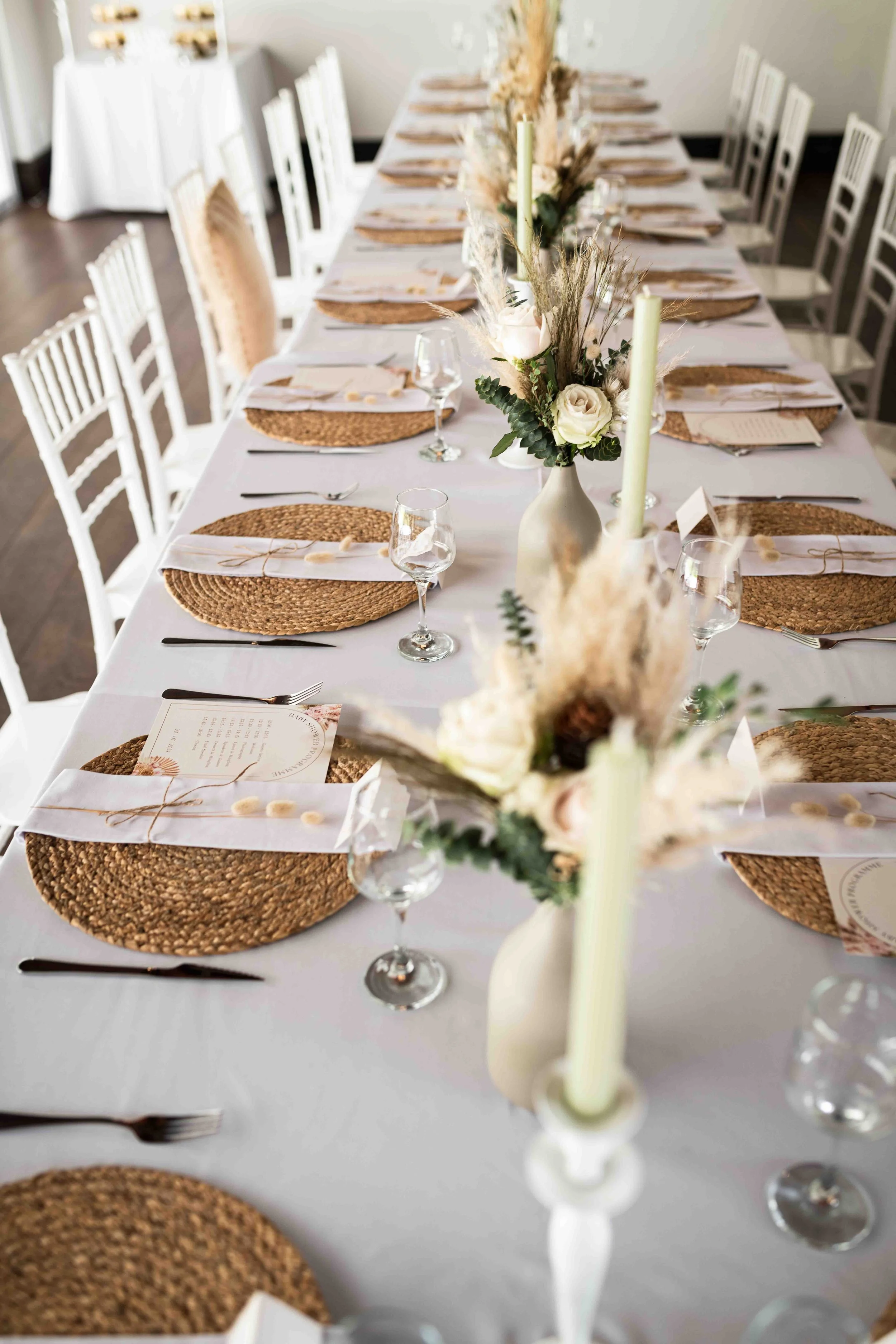 A photograph taken during a baby shower event showing a long table set with woven round placemats, white cloth napkins , wine glasses, and small menu cards, decorated with cream-coloured vases filled with white roses and dried grasses, and candles.