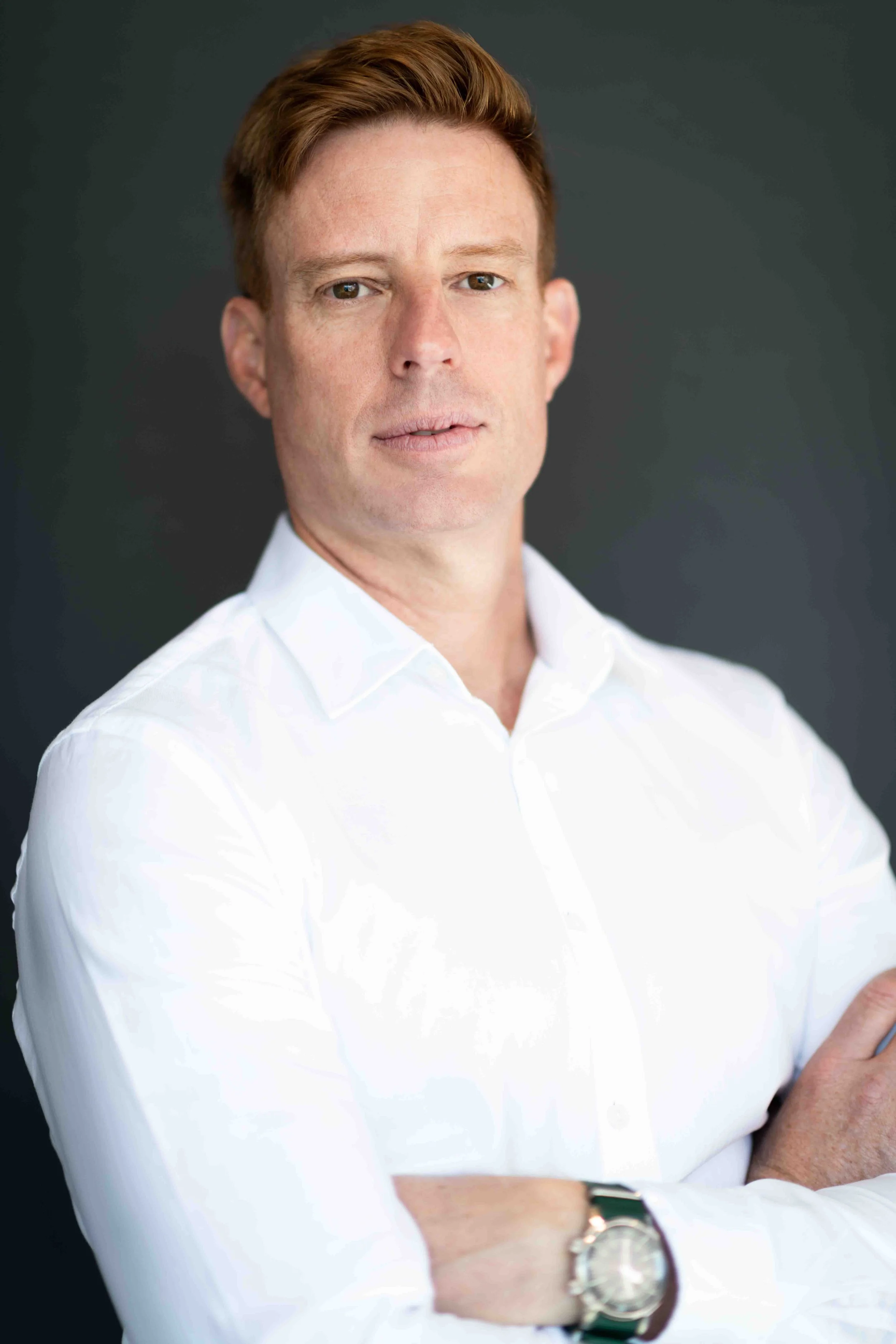 A corporate professional headshot taken in Cape Town of a man, wearing a white shirt and watch, posing with arms crossed against a dark background.