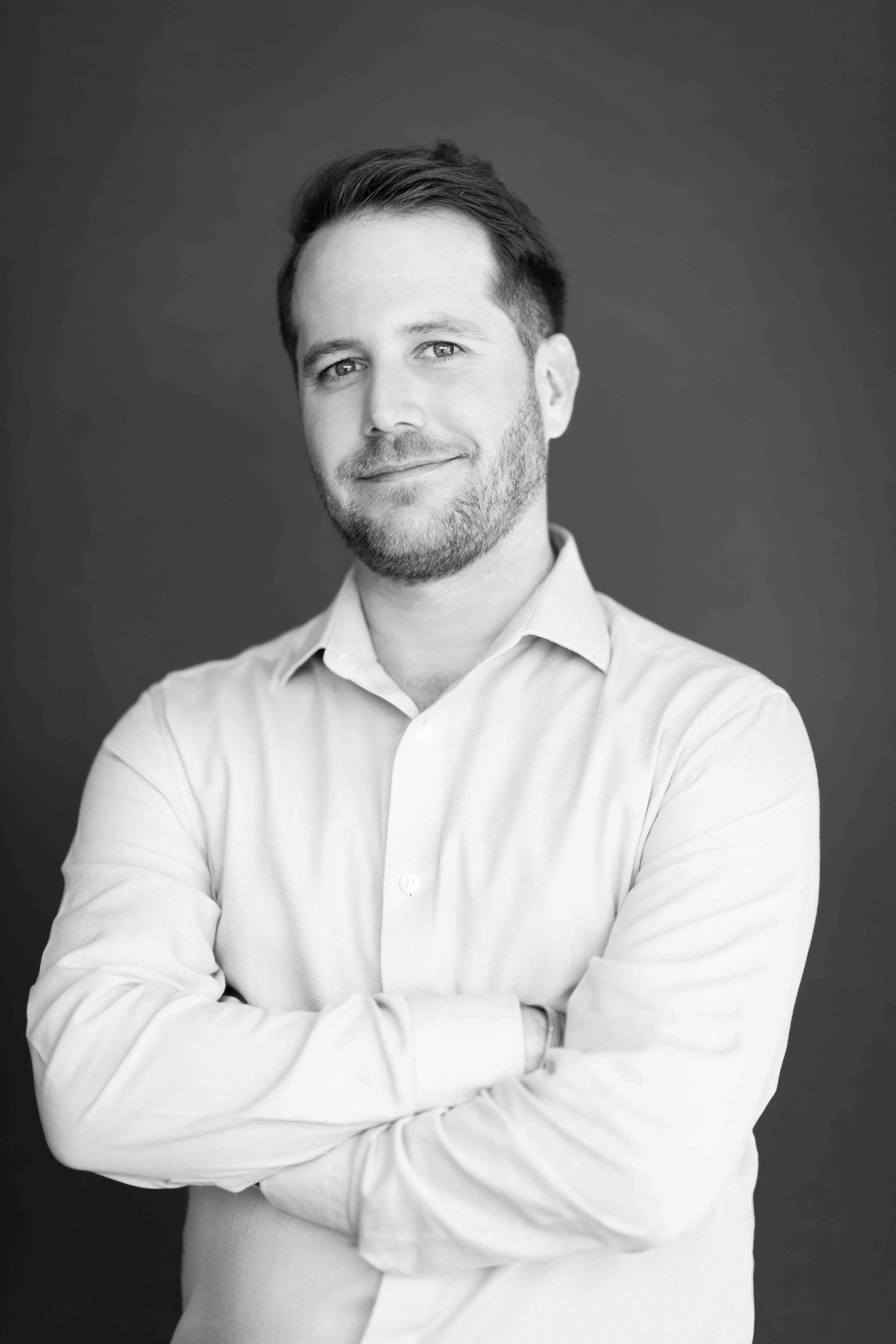 A corporate professional headshot taken in Cape Town in black and white of a man with short hair and a beard, wearing a light-colored button-up shirt, smiling with arms crossed in front of a dark background.
