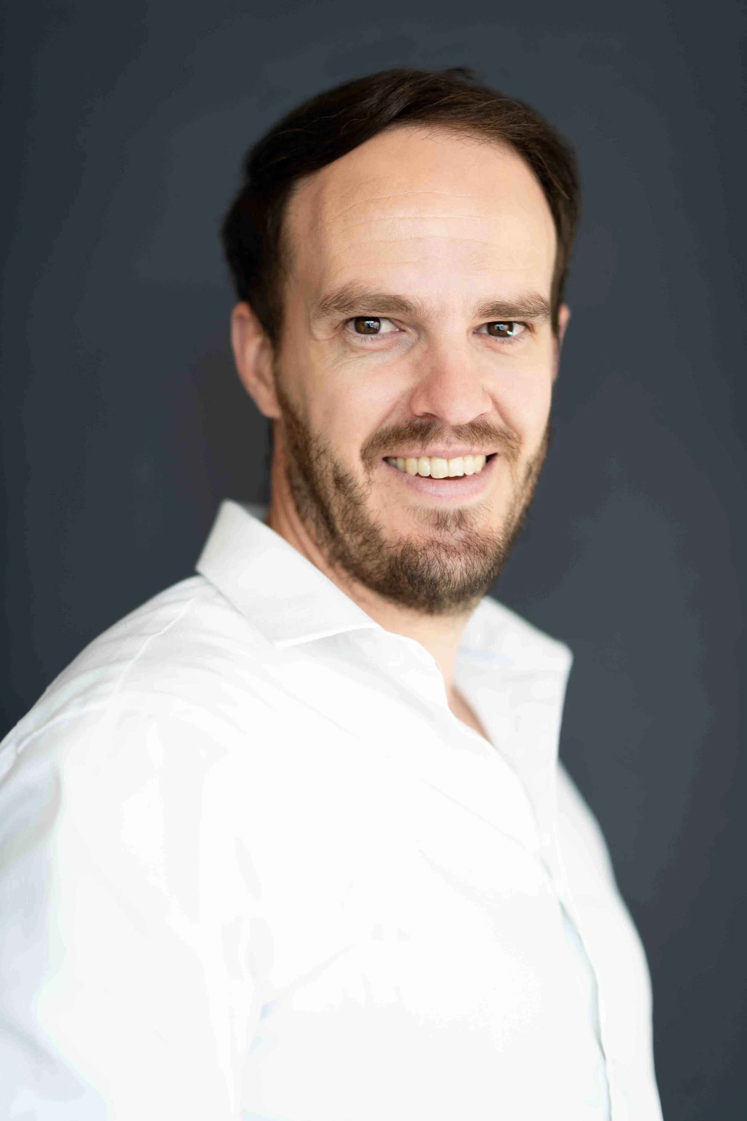 A corporate professional headshot taken in Cape Town of a man with brown hair and a beard smiling, wearing a white shirt, against a dark background.