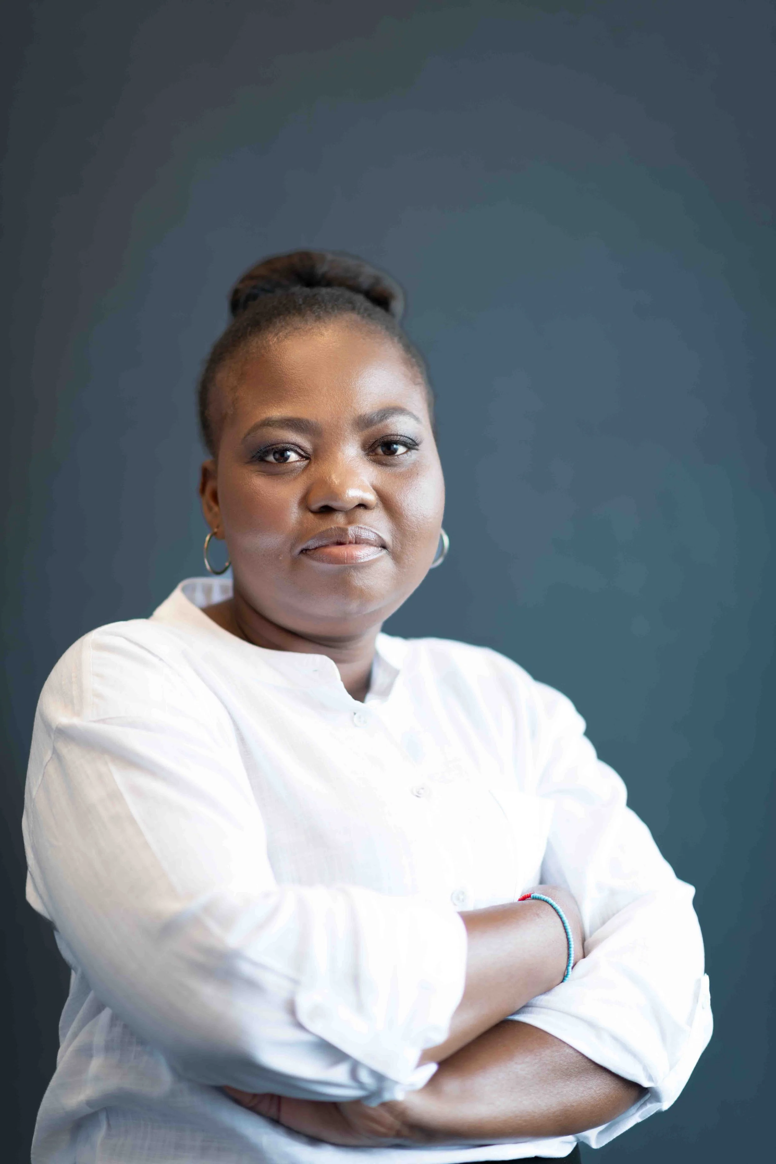 A corporate professional headshot taken in Johannesburg of a confident woman with arms crossed, wearing a white shirt and hoop earrings, standing against a dark background.