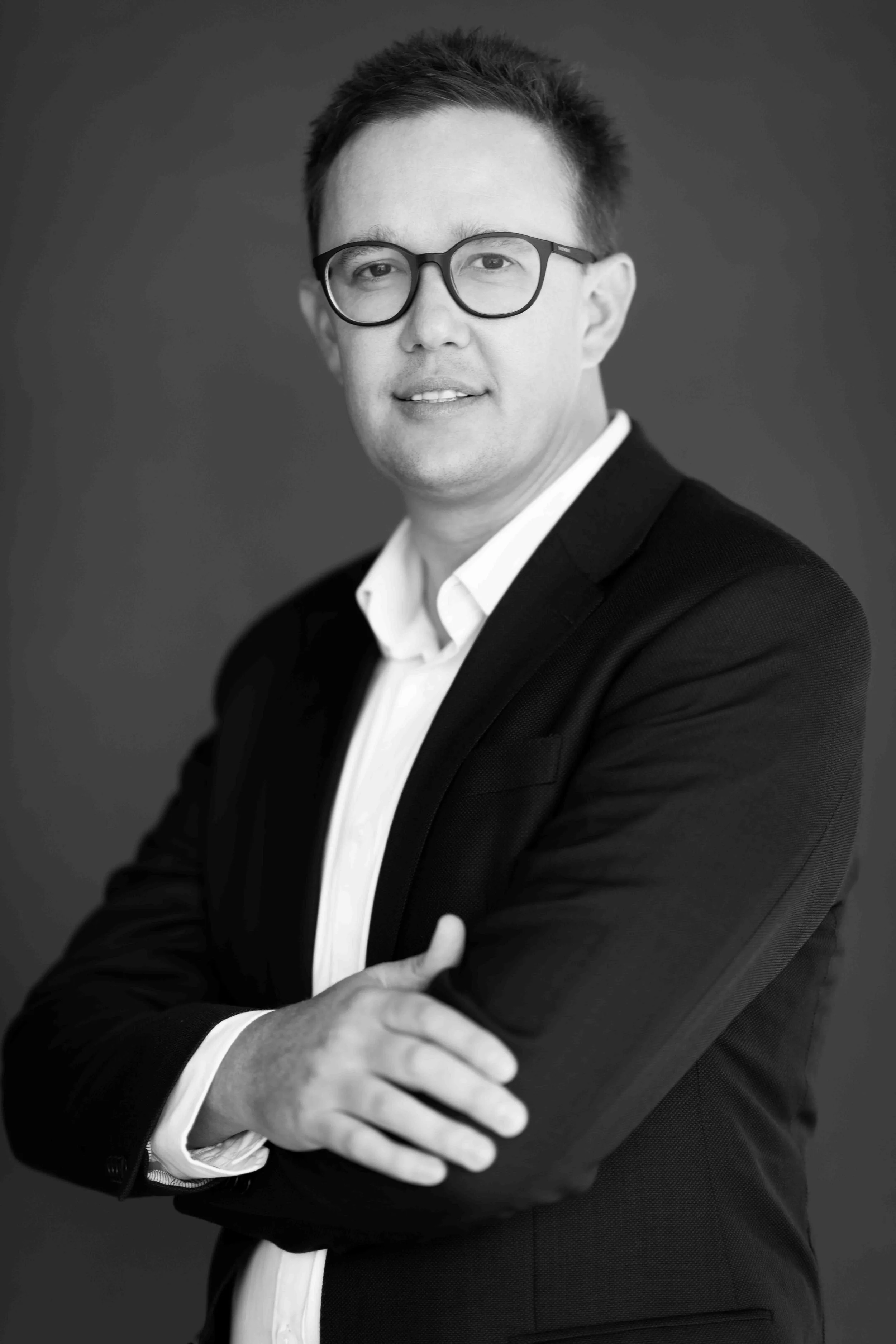 A corporate professional headshot taken in Cape Town in black and white of a man wearing glasses, a dark suit, and a white dress shirt, with crossed arms, against a plain background.