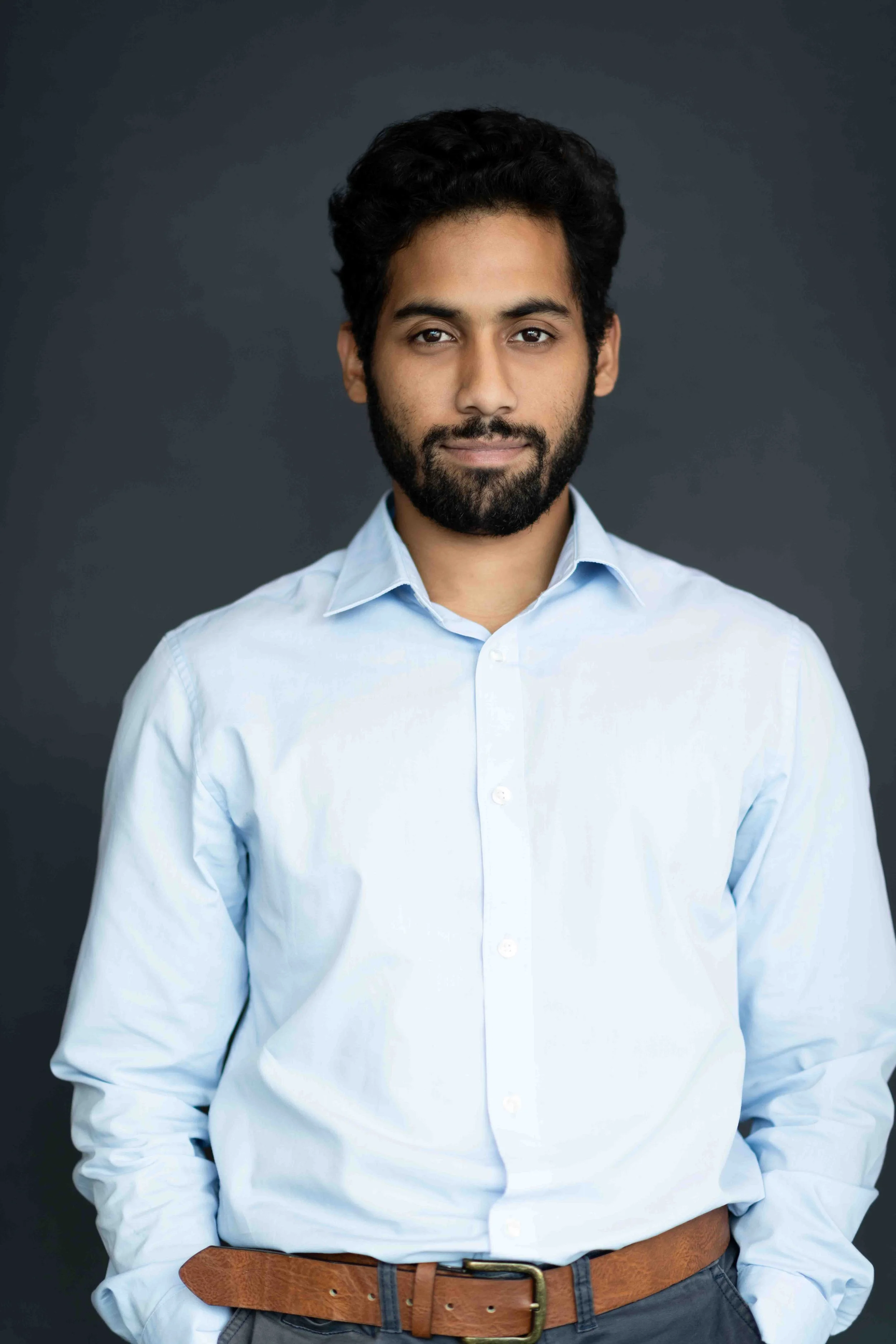 A corporate professional headshot taken in Cape Town of a man with dark hair, beard, and light blue dress shirt standing against a dark grey background.