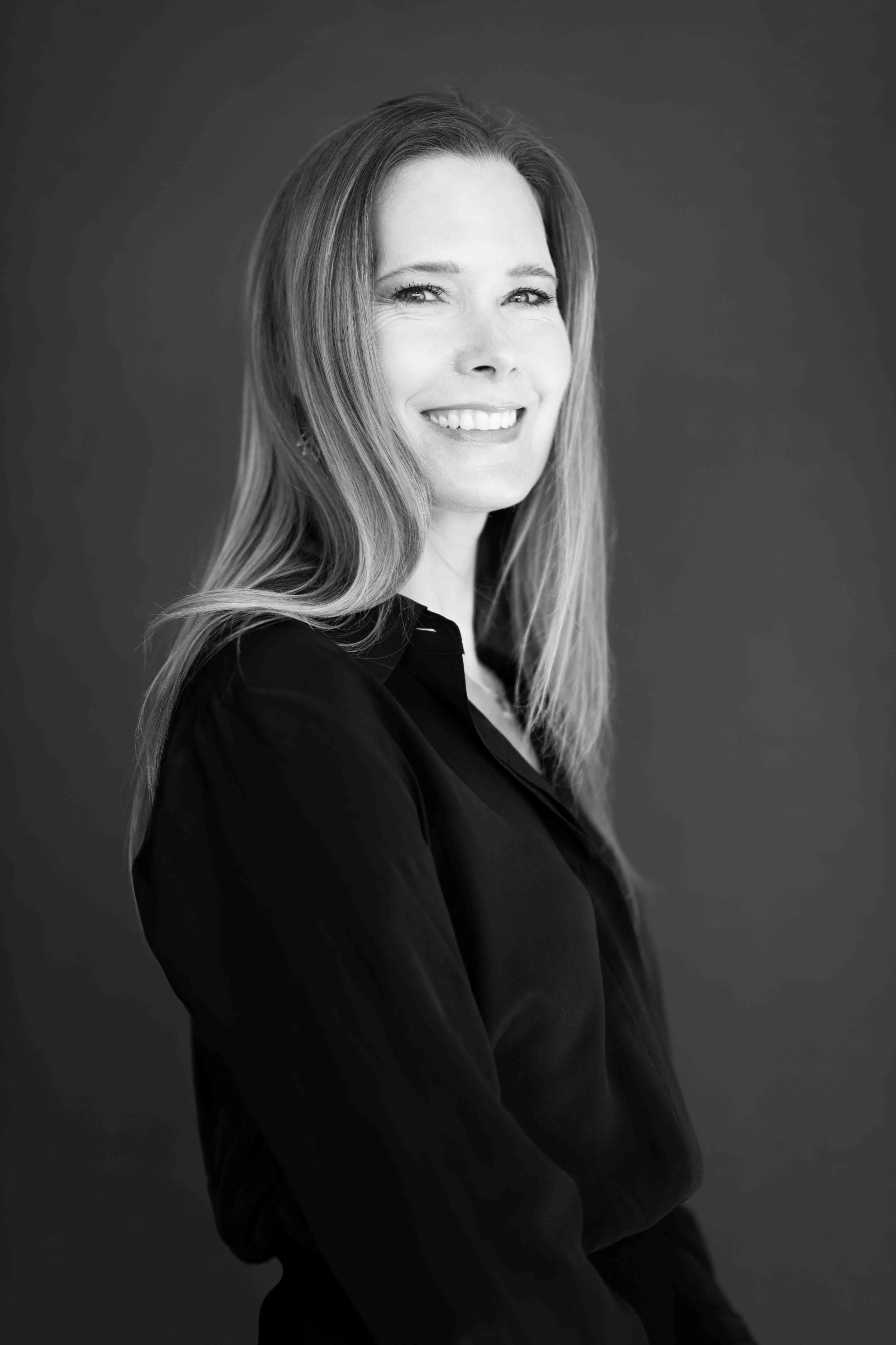 A corporate professional headshot taken in Cape Town in black and white of a woman with long hair, smiling, wearing a dark blouse, against a plain background.
