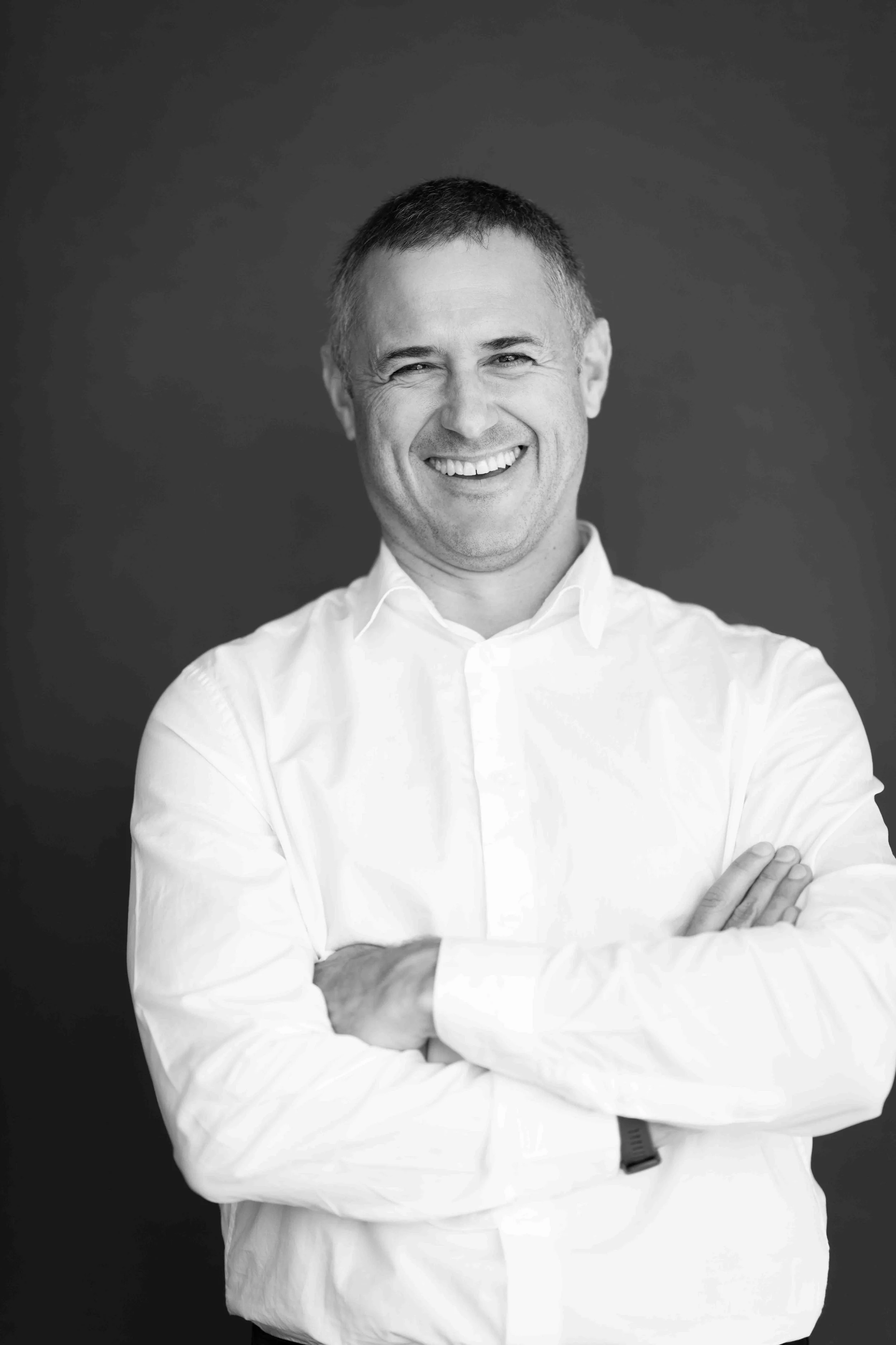 A corporate professional headshot taken in Cape Town in black and white of a smiling man with short hair, wearing a white collared shirt, standing with arms crossed against a dark background.