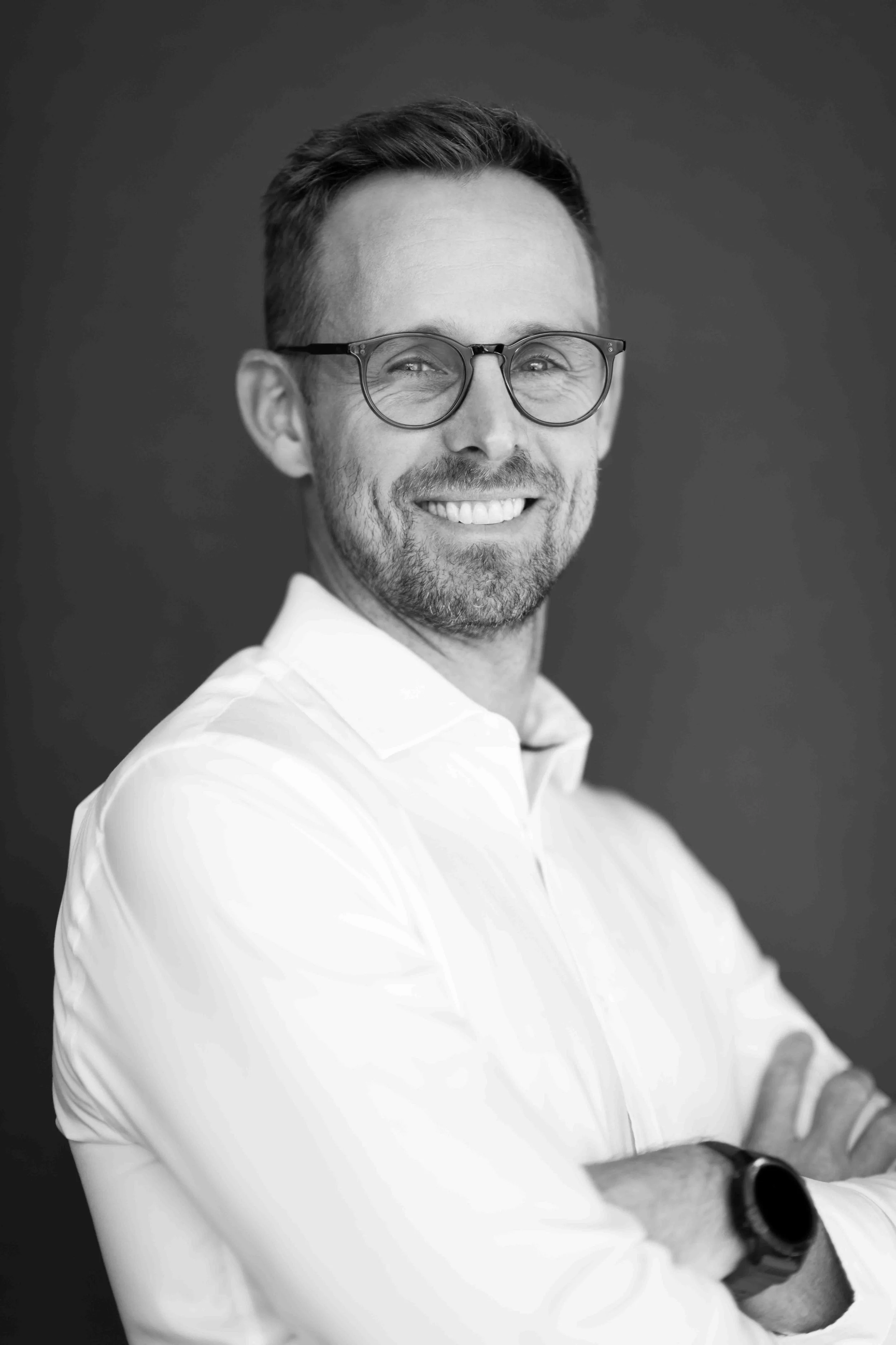 A corporate professional headshot taken in Cape Town in black and white of a smiling man with glasses wearing a white shirt, arms crossed, against a dark background.