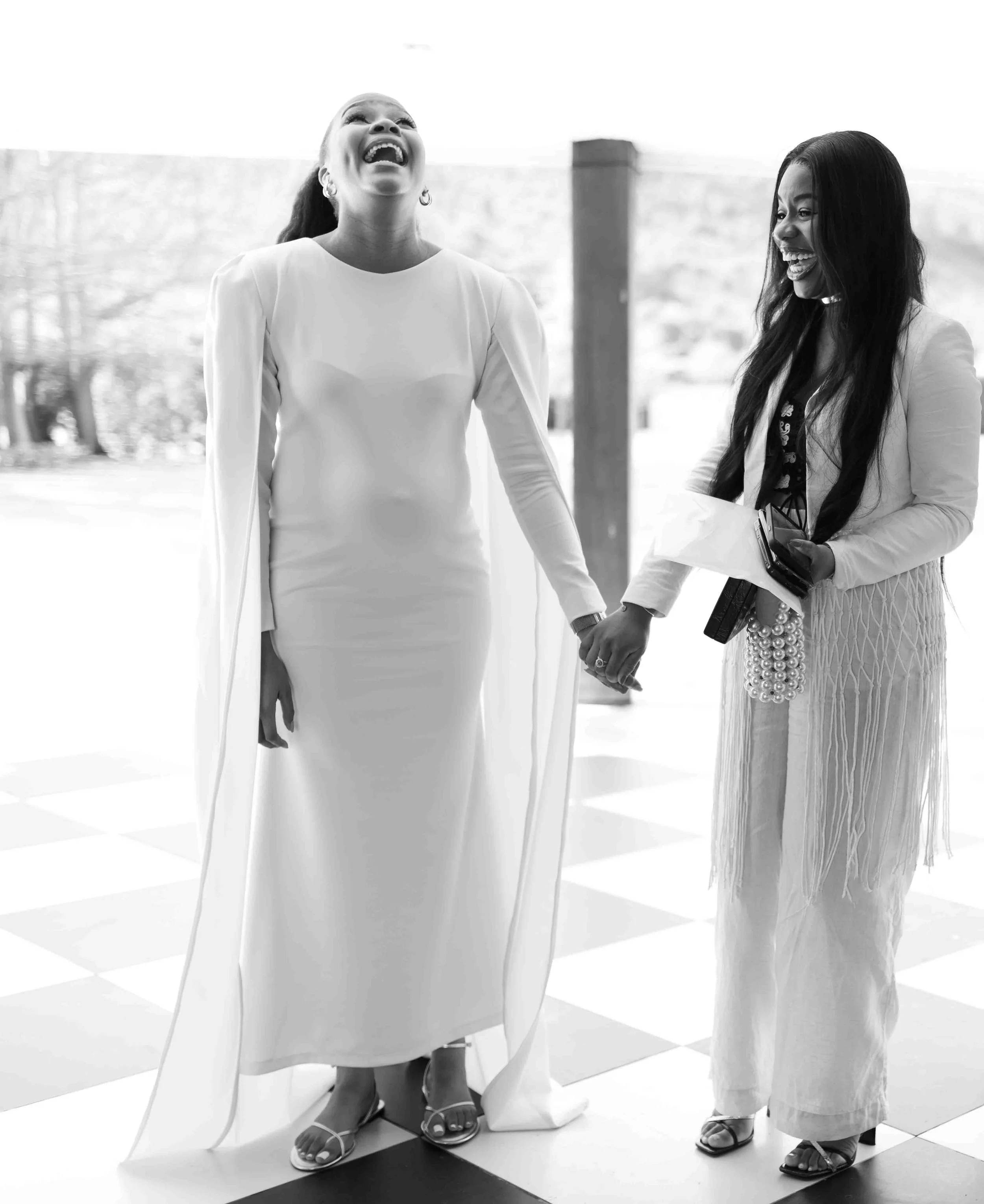 A photograph taken during a baby shower event showing two women smiling and holding hands, one in a white dress with cape and the other in stylish clothing, standing on a checkered floor in an indoor setting.